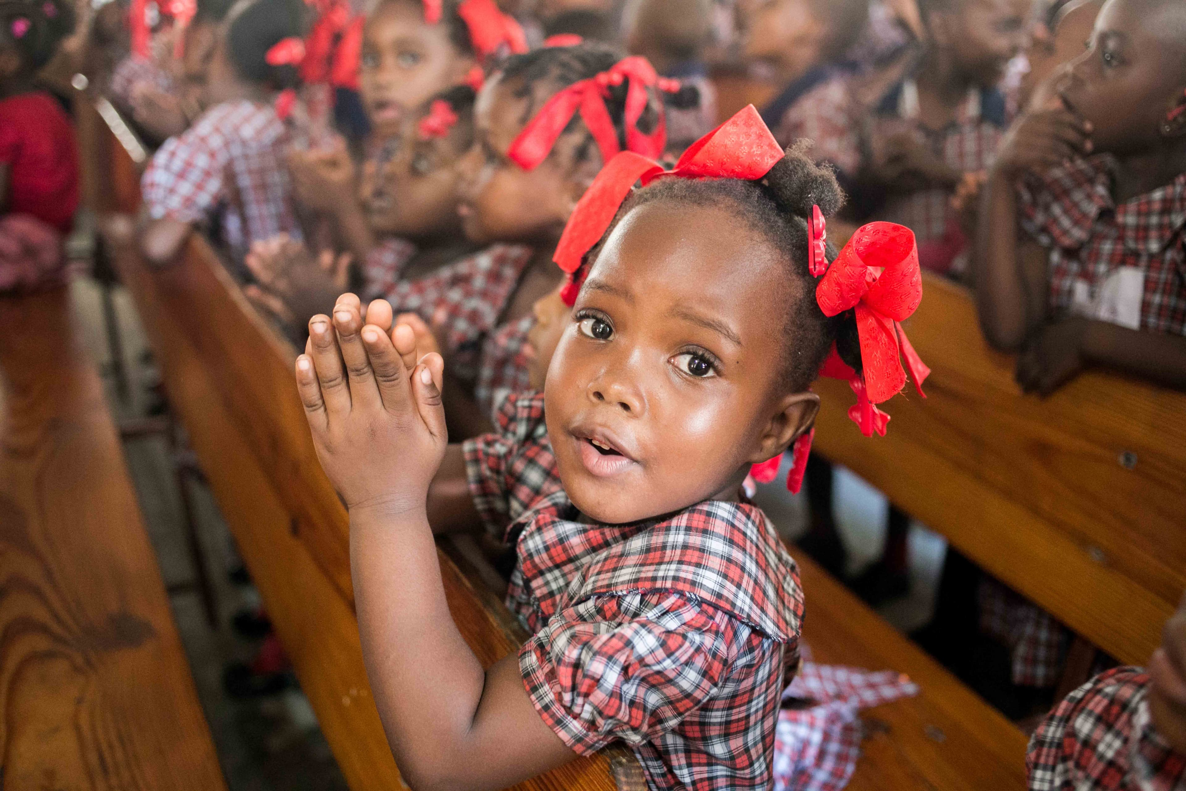A little girl in a red plaid uniform and red hair ribbons holds her palms together in prayer