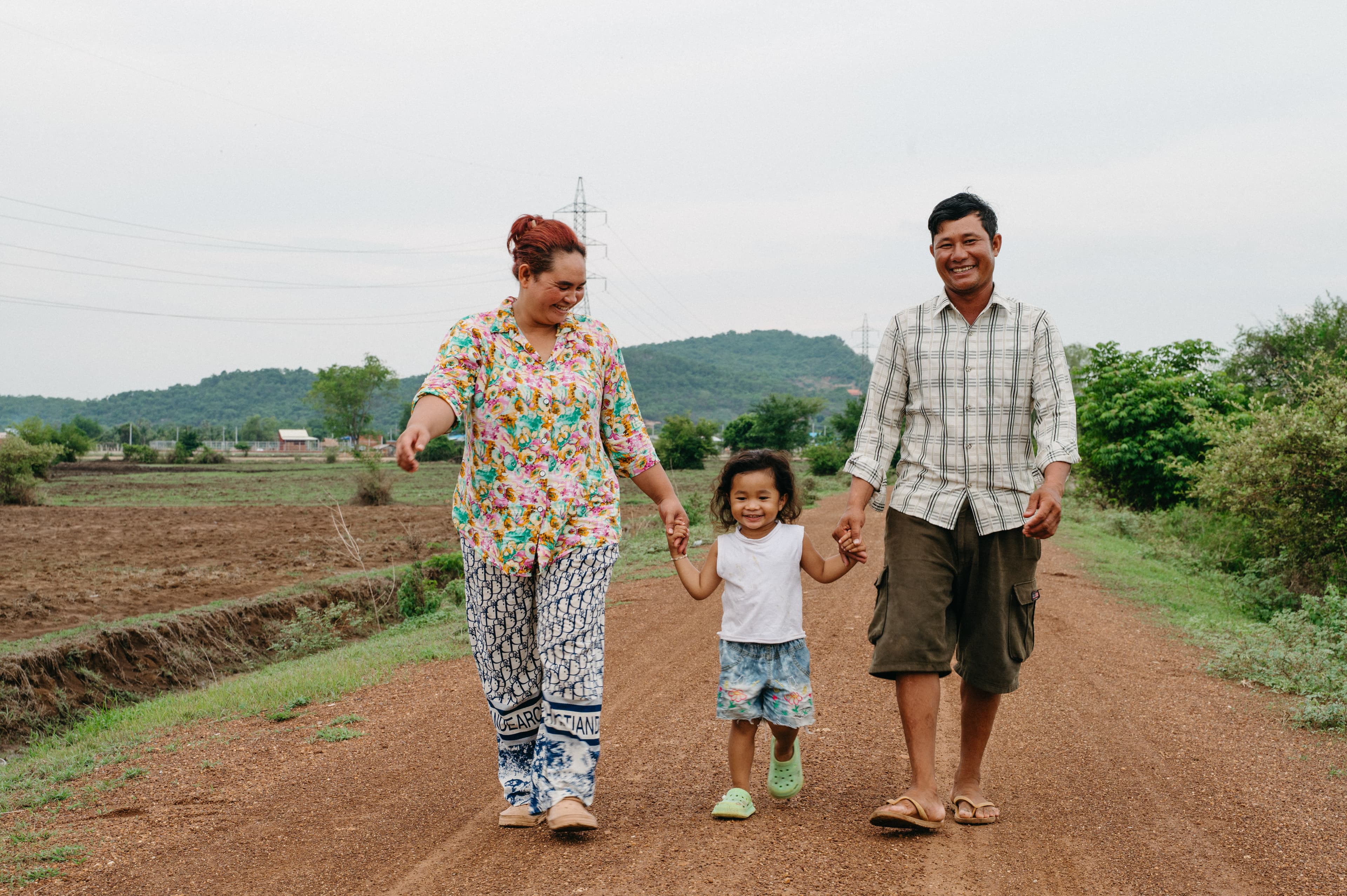 A young girl walks between her parents on a dirt road holding their hands as they all smile at the camera and behind them is a mountain.