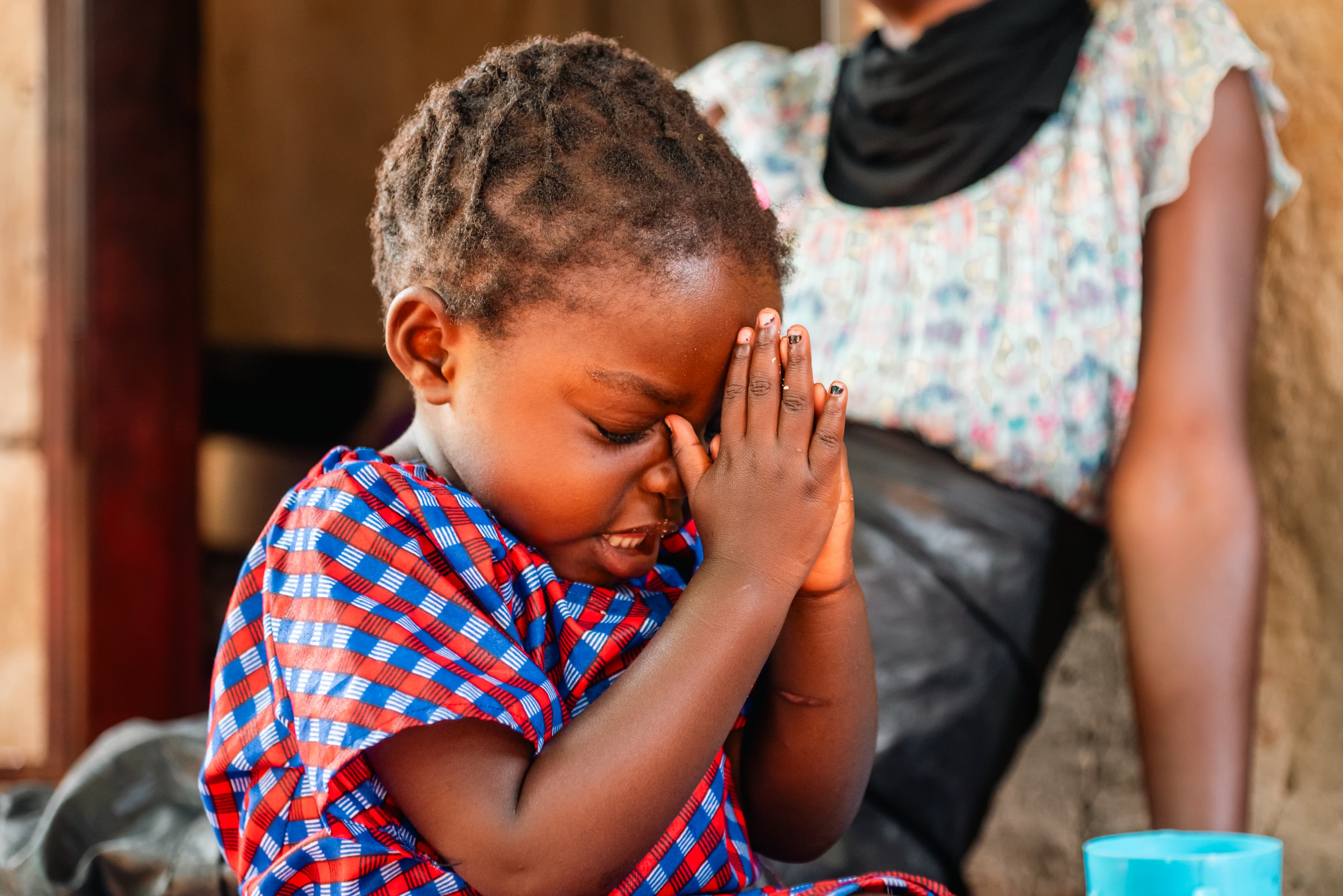 A young girl, wearing a blue and red printed dress, closes her eyes and holds her hands together to pray.