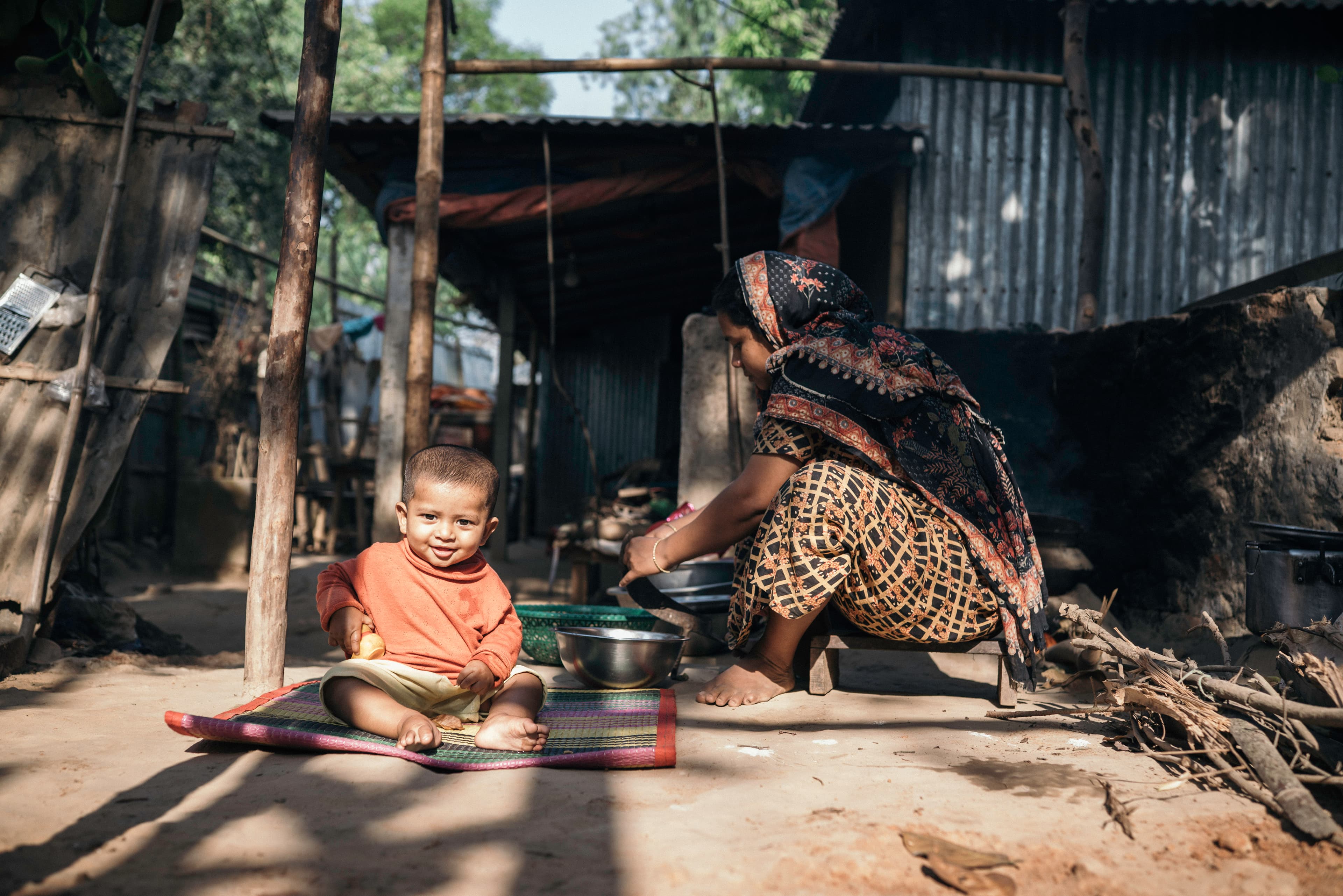 A toddler girl sits on a mat and smiles with her mother cooking behind her.