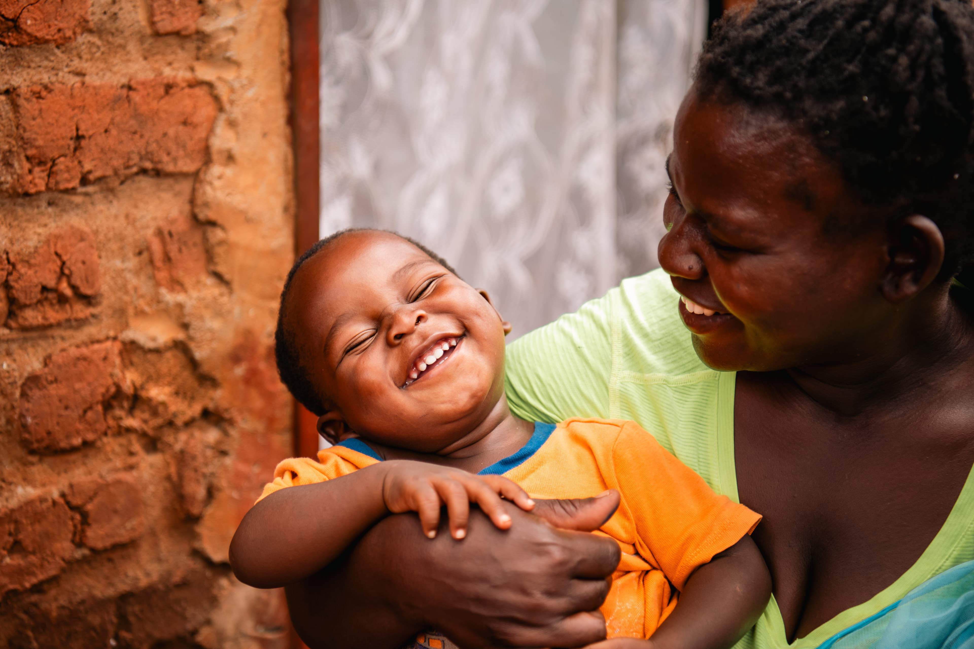 A smiling woman in a yellow shirt stares down at her laughing 2-year-old she is holding in her arms.