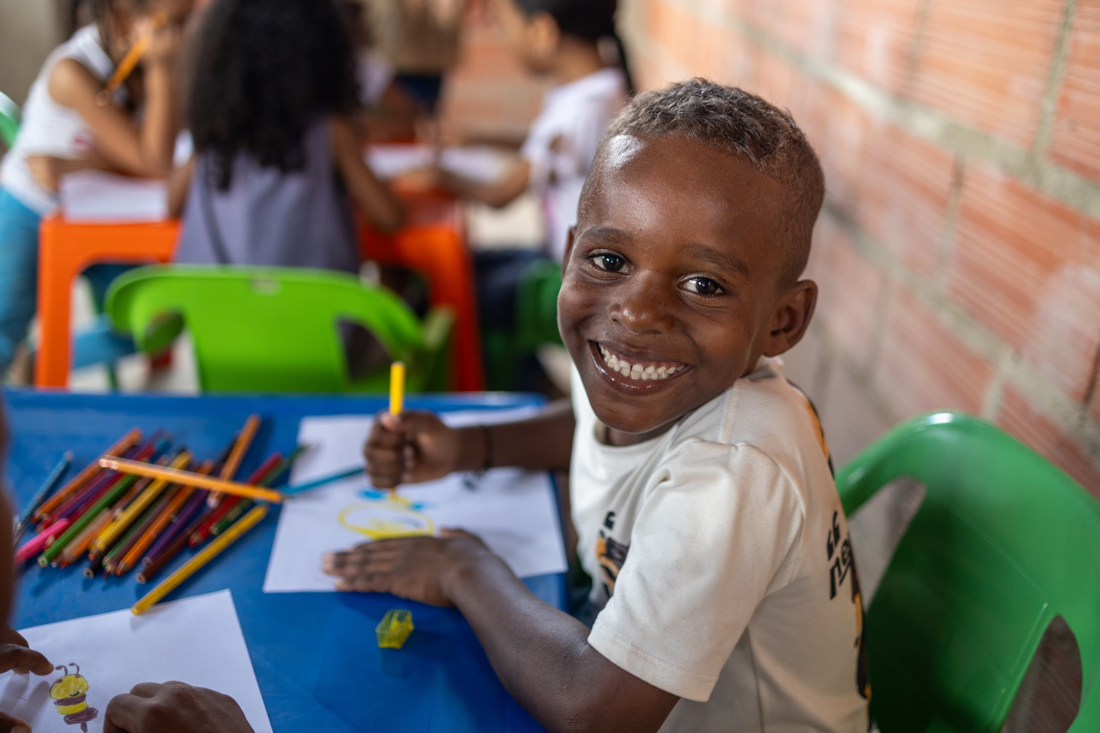 Young, Colombian boy wearing a white T-shirt smiles while coloring at a blue table.