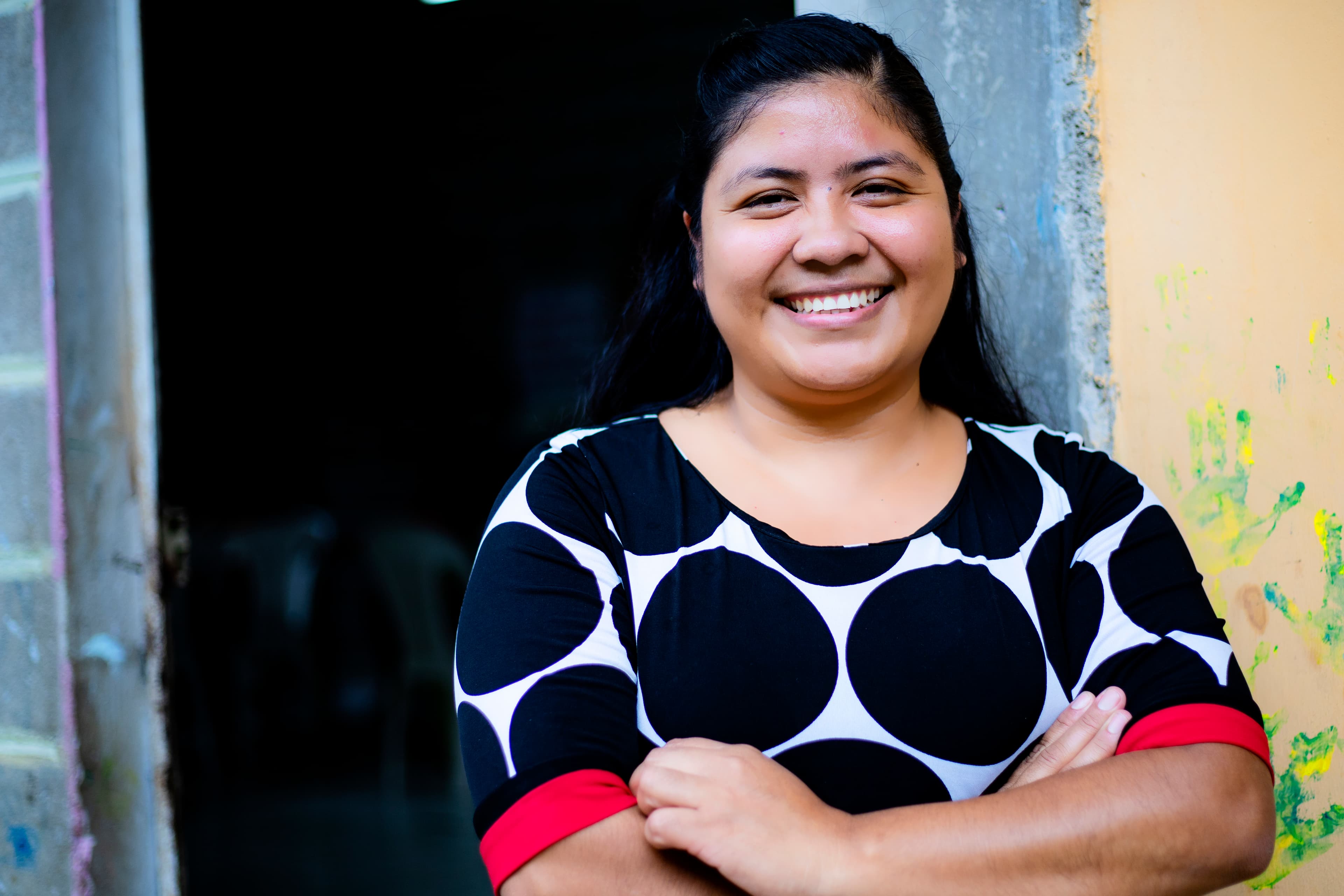 A young woman smiles at the camera with her arms crossed. She is wearing a black and white polka dot top and leaning on a painted concrete wall.