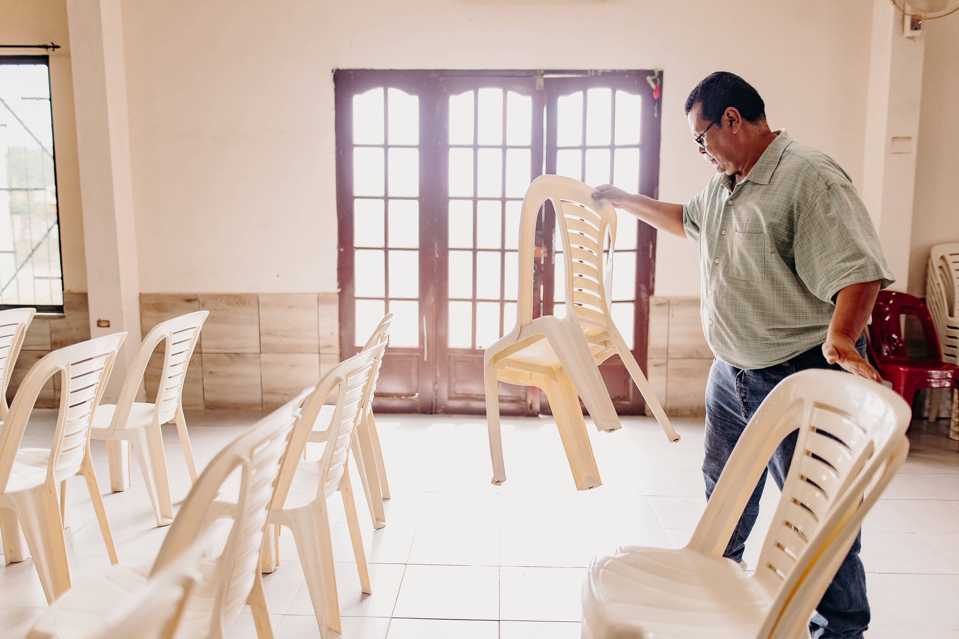 An older man works to put out plastic chairs in front of a bright door.