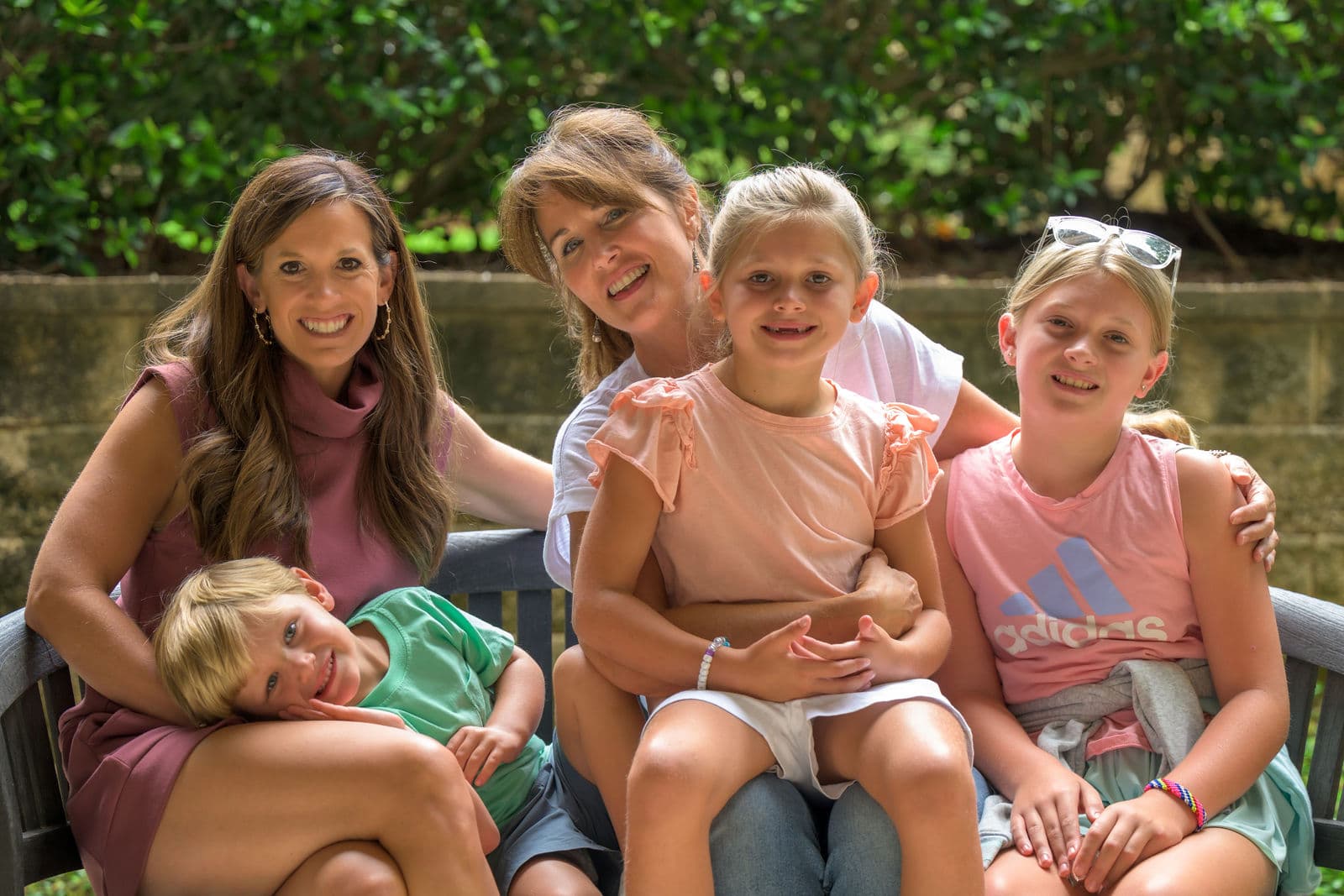 Two women and three children sit on a grey bench outside, in front of a stone wall and plants.