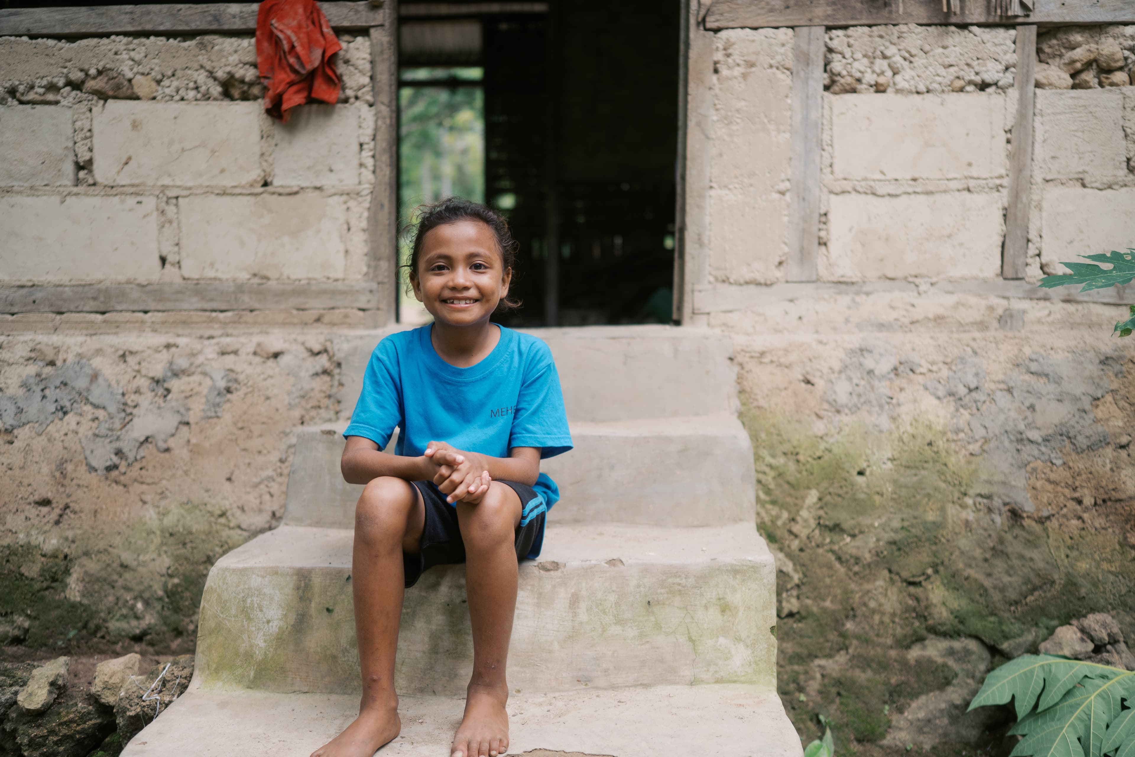 A young girl is sitting on the staircase outside her home with her hands clasped together in her lap.