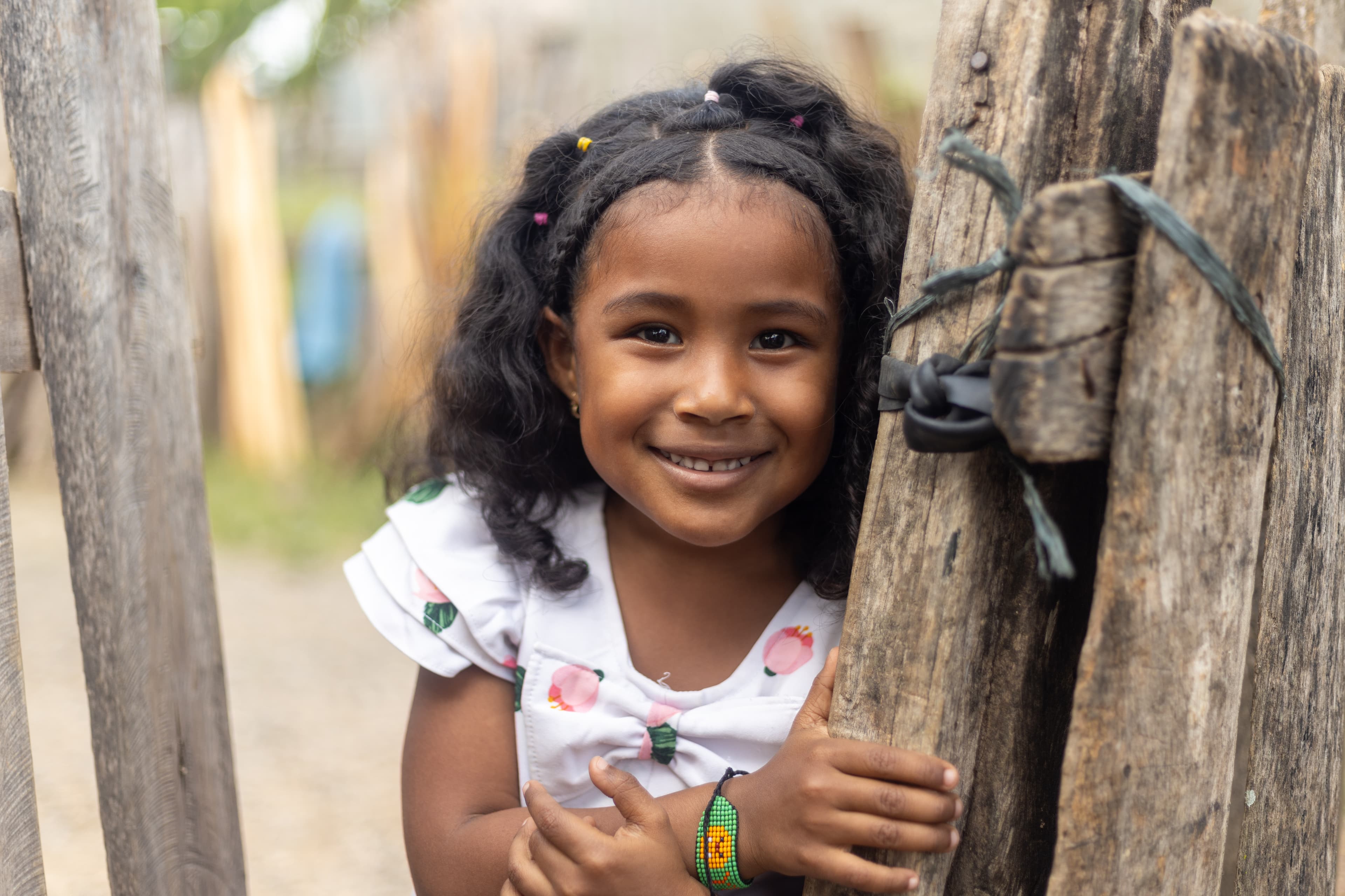 A little girl leans against a wood fence and smiles.