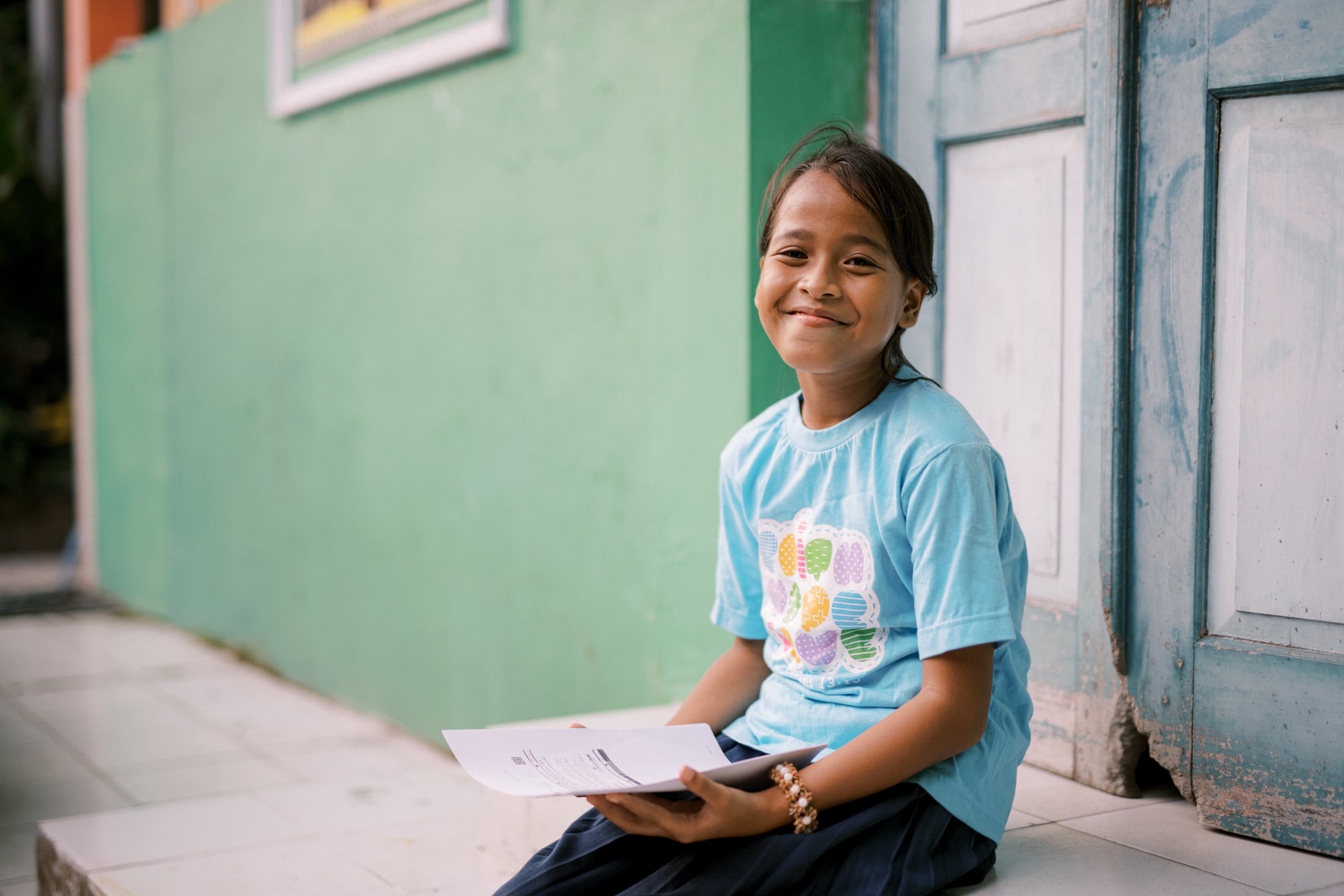 A young girl sits outside a home holding a sponsor letter and smiling.