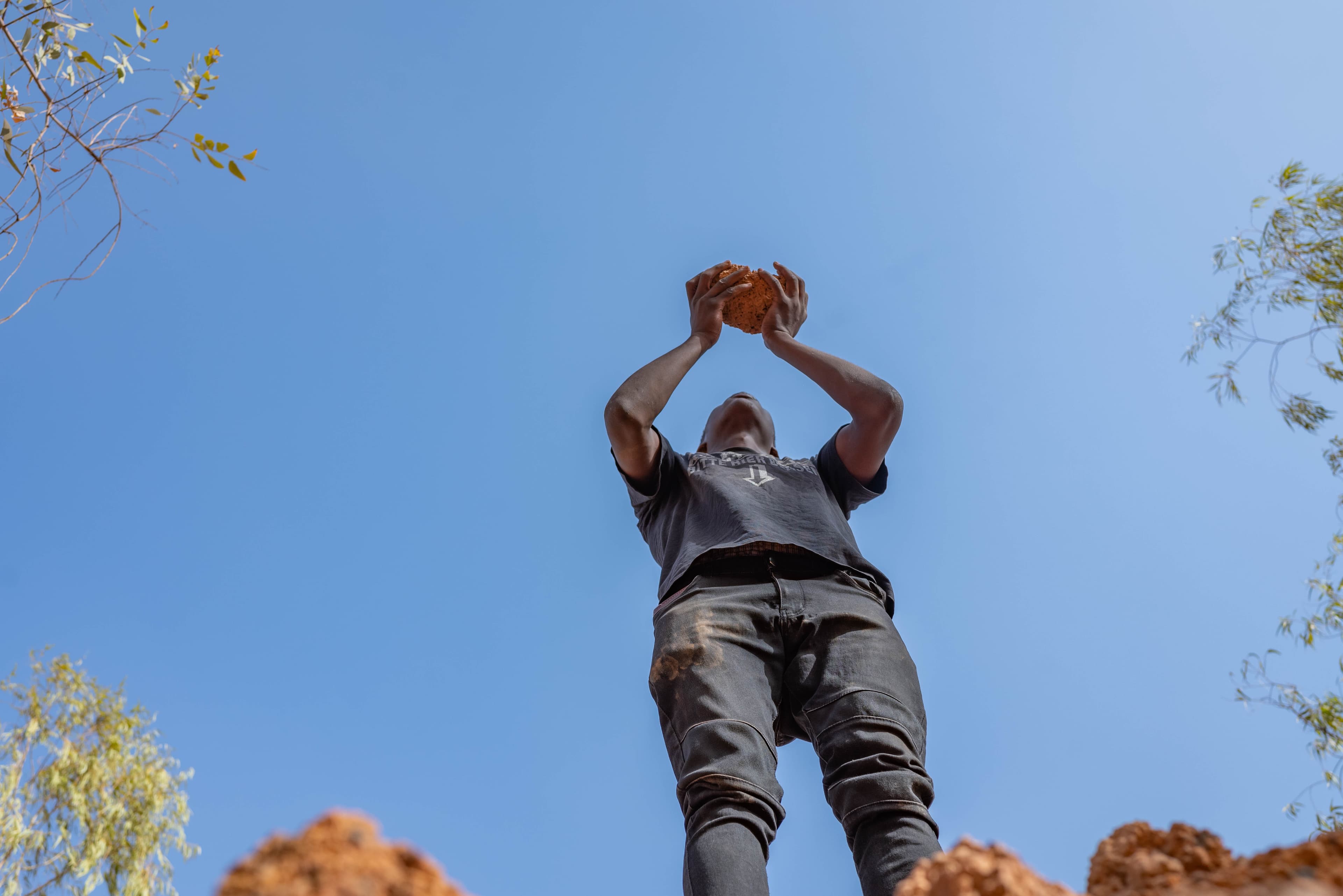 A teen African boy stands and lifts a rock into the air.