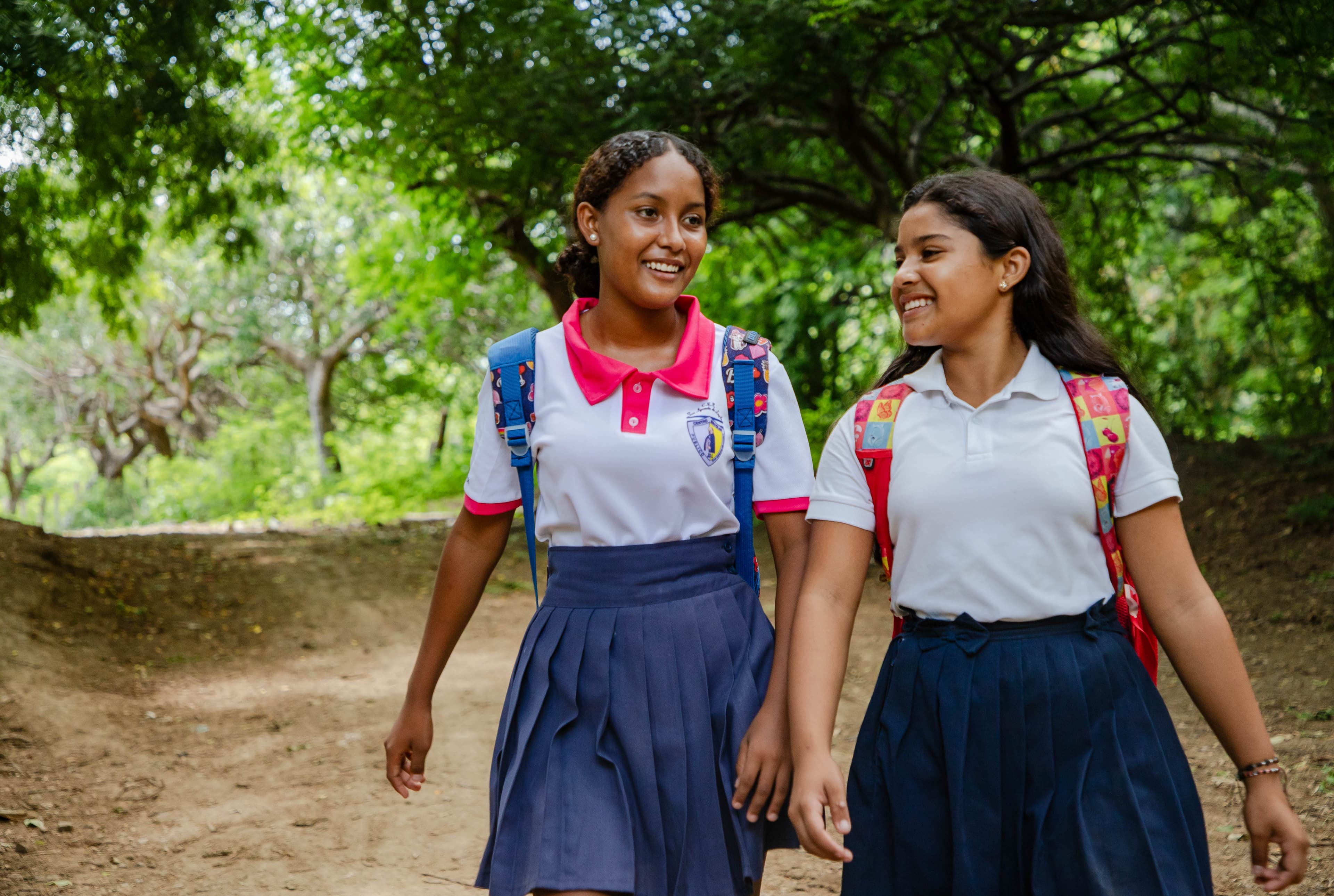Two girls wearing backpacks and school uniforms walk and talk together.