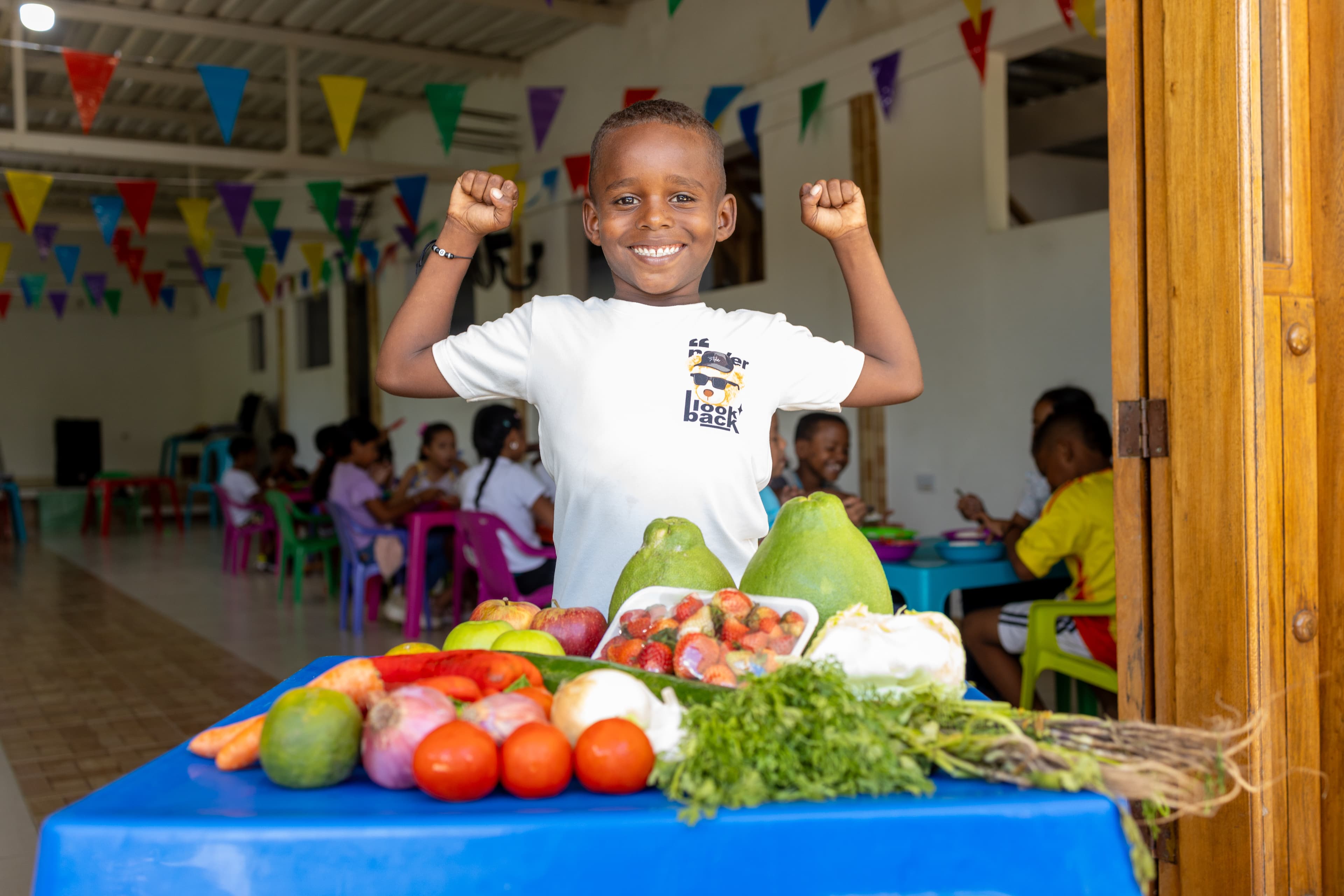 A young boy flexes his muscles and smiles in front of a table of vegetables.