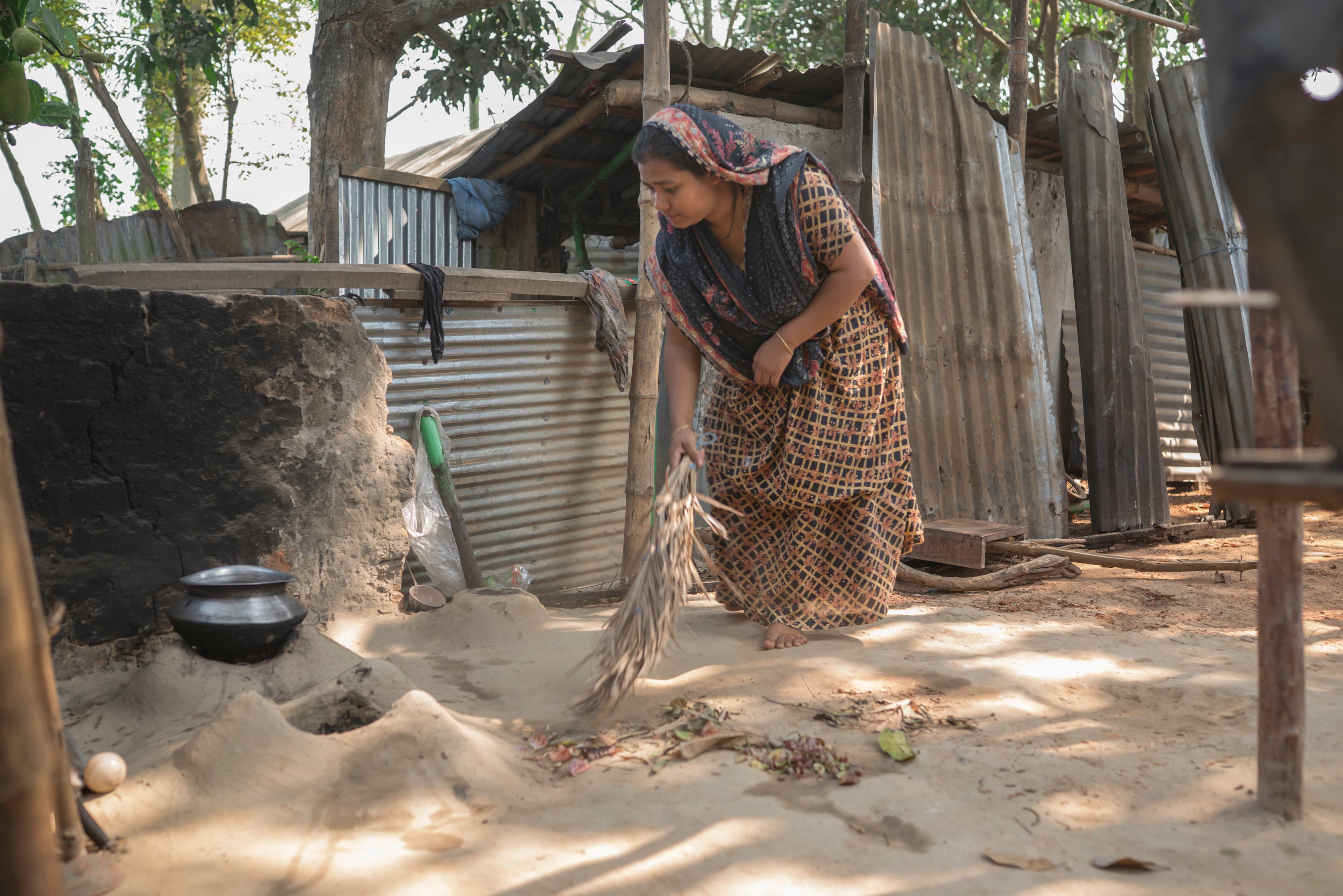 A woman wearing a headscarf and dress sweeps the outside of her home with branches.