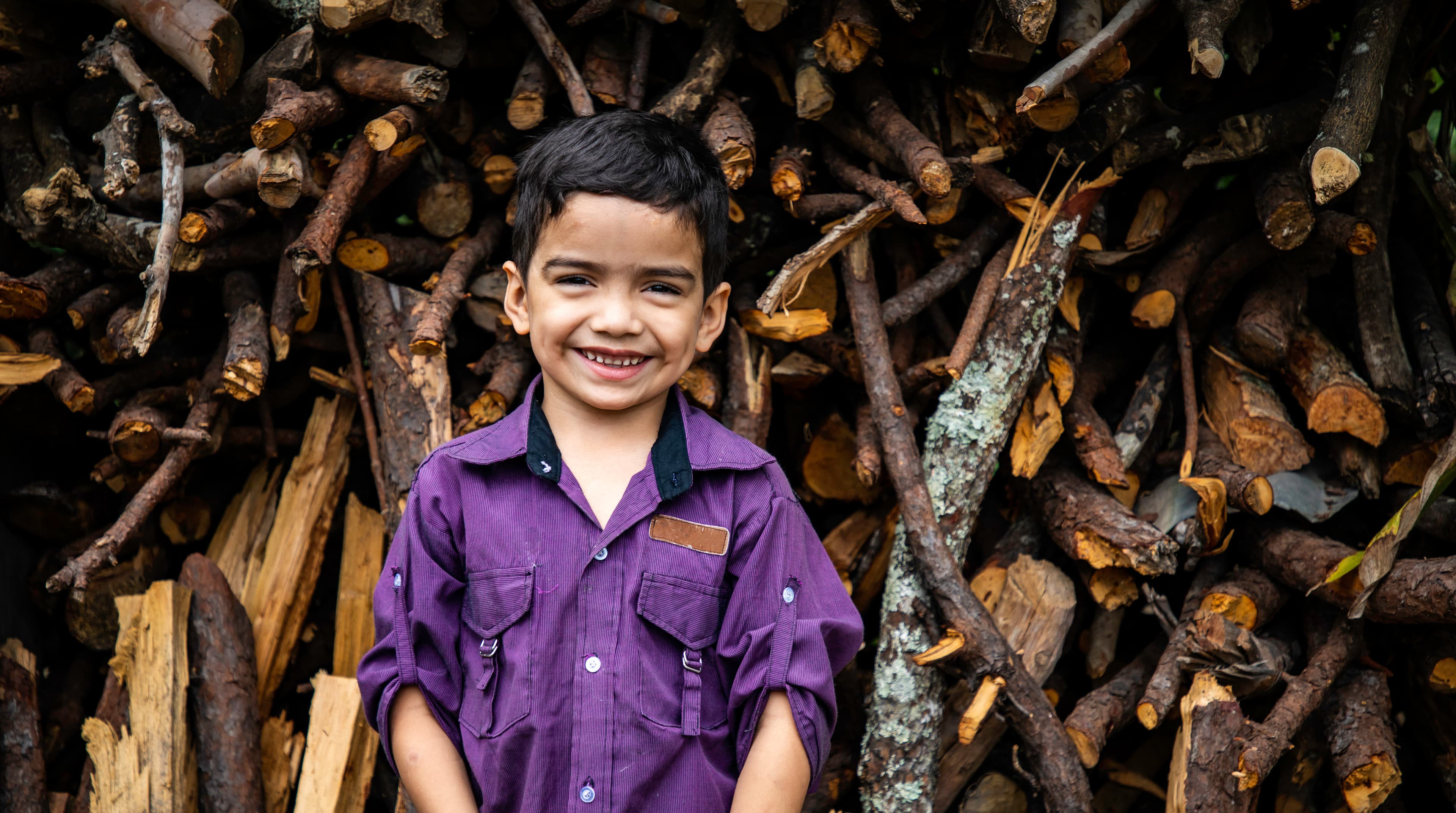A young boy is smiling and standing in front of a pile of firewood.