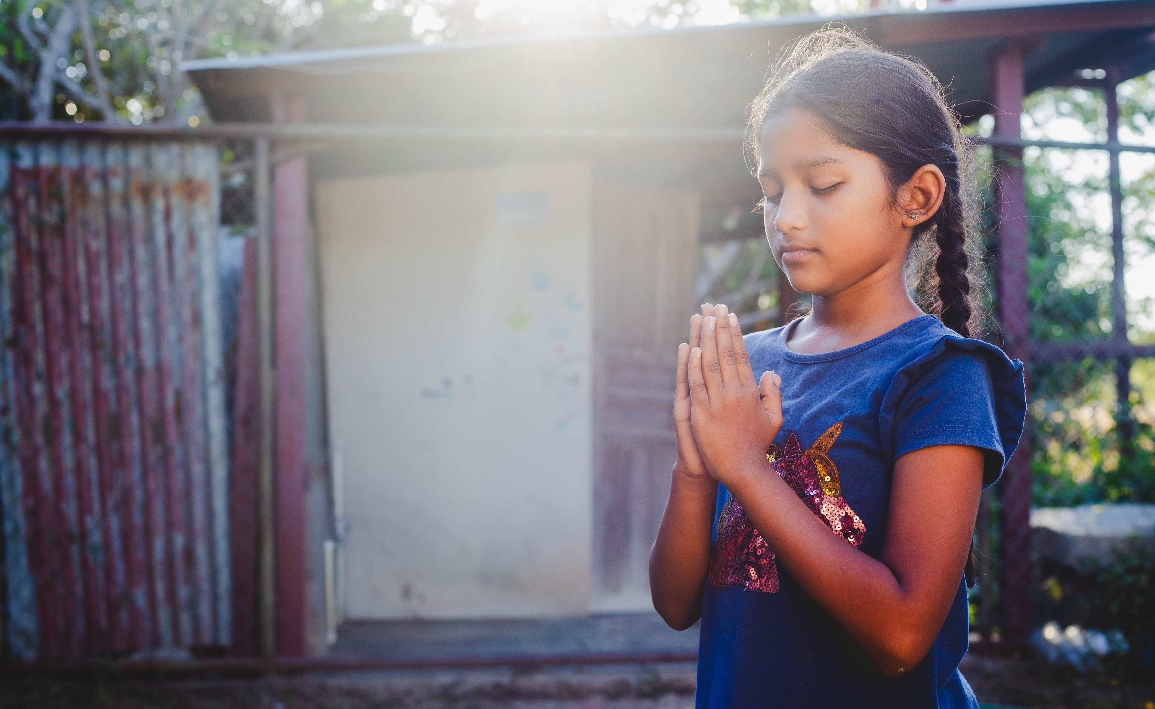 A girl in a blue shirt stands outside, praying with her eyes closed and hands folded.