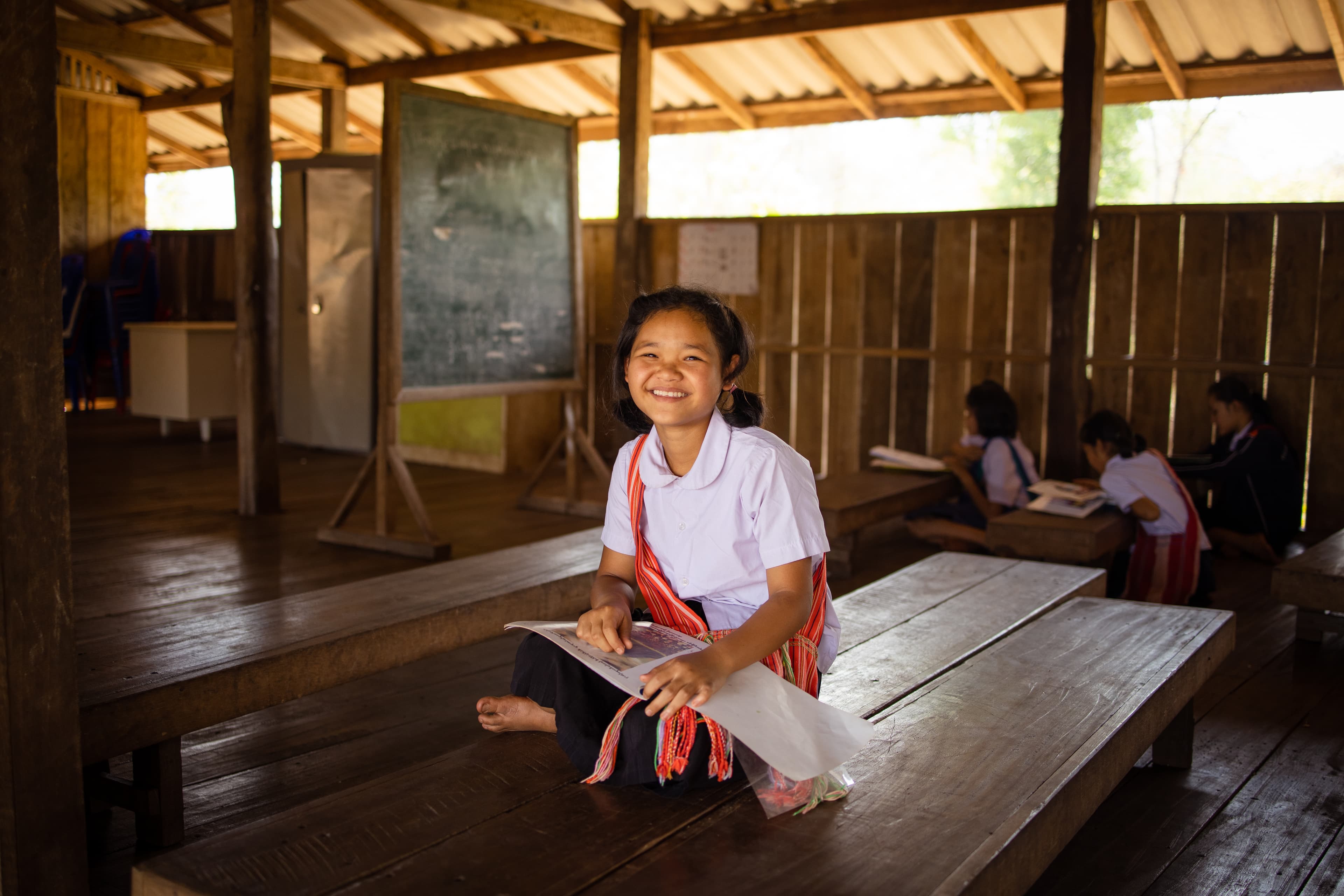 A young Thai girl sits on a wooden bench holding a book and smiling.