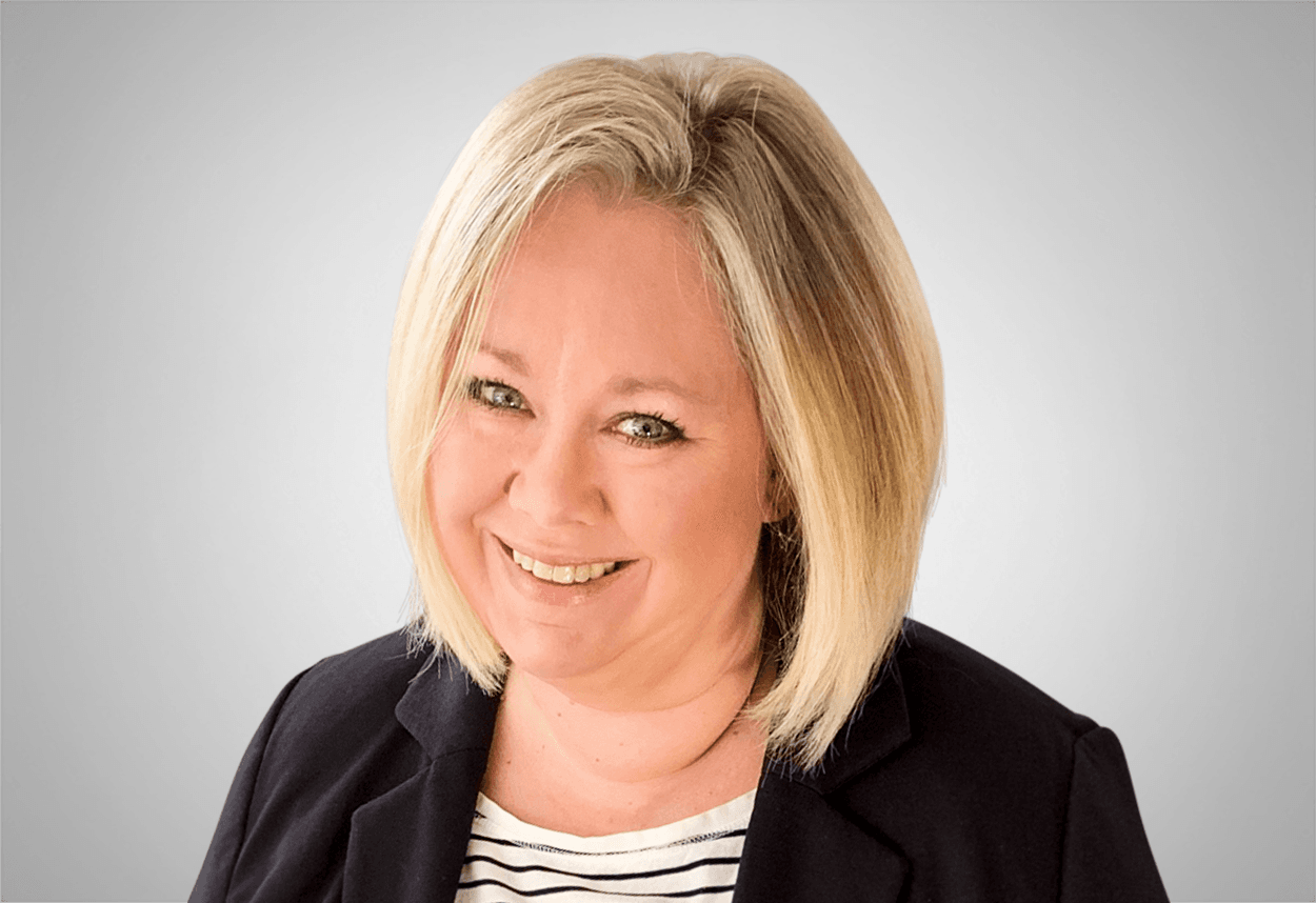 Danika Mitcham is in a black and white striped shirt with a black blazer is smiling, with a grey background behind her.