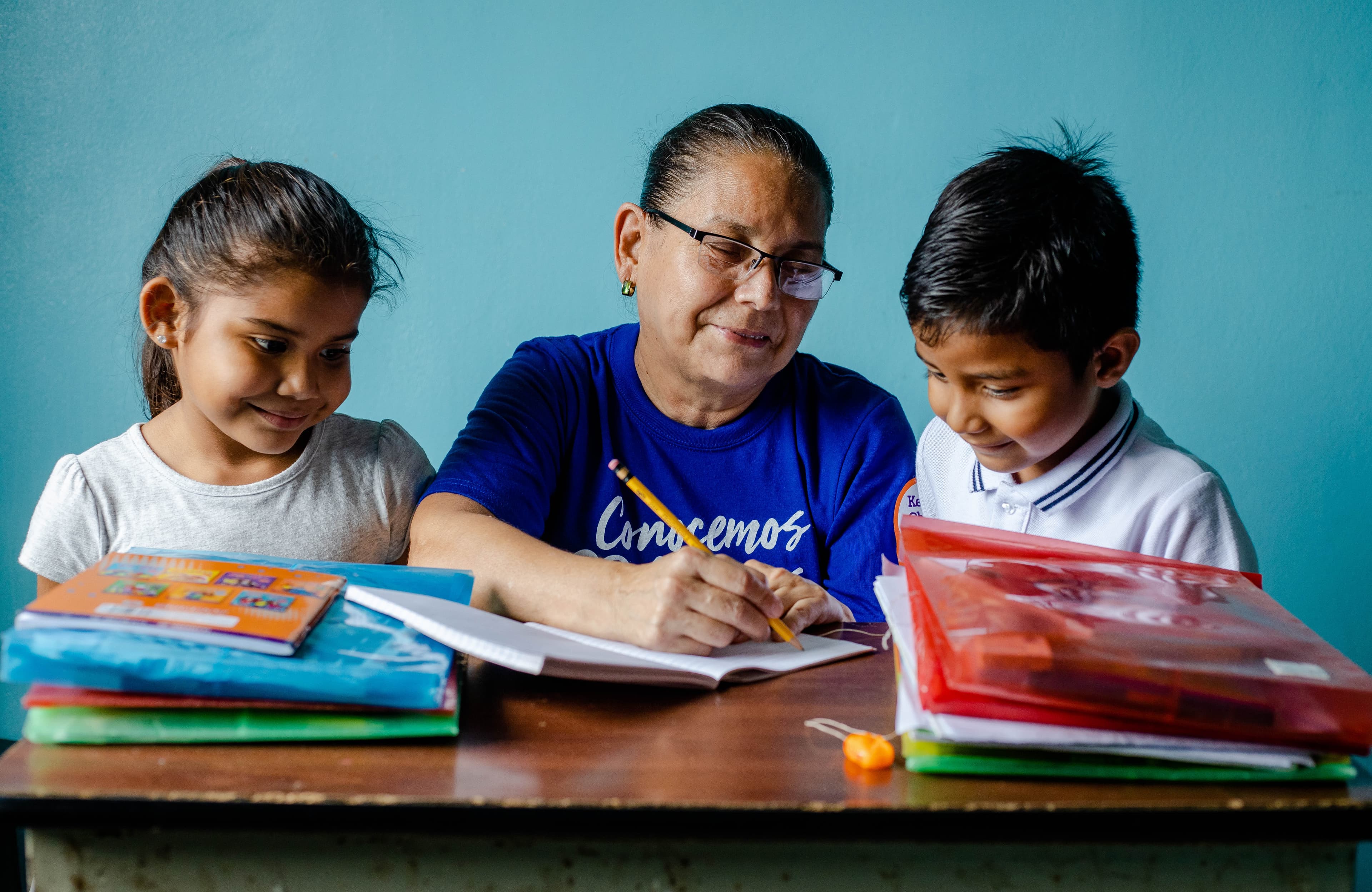 A woman is sitting at a desk with two children who are looking at what she is writing in a notebook.