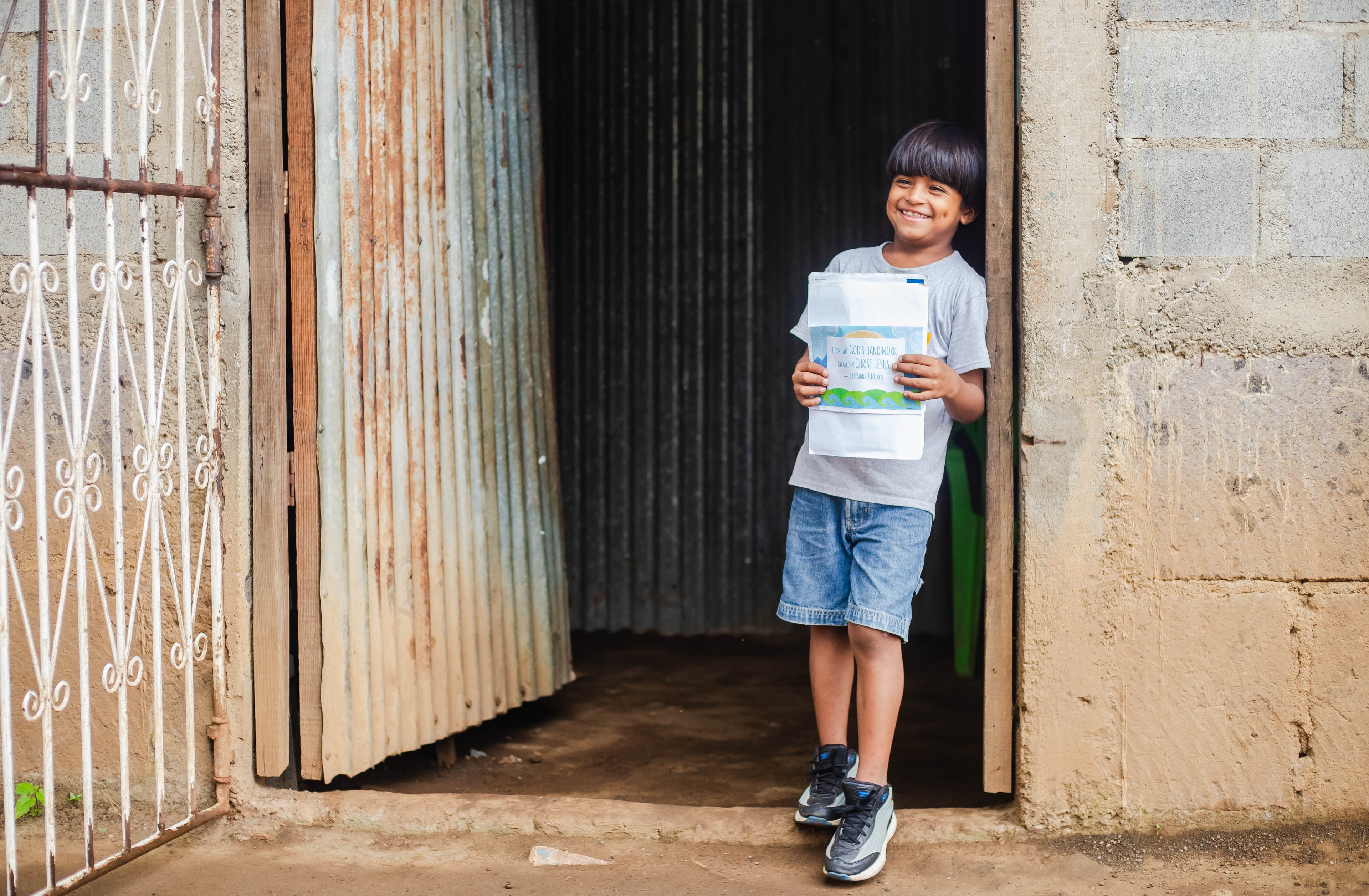 A boy wearing a gray T-shirt and jean shorts stands in the doorway of a building holding a letter and smiling.