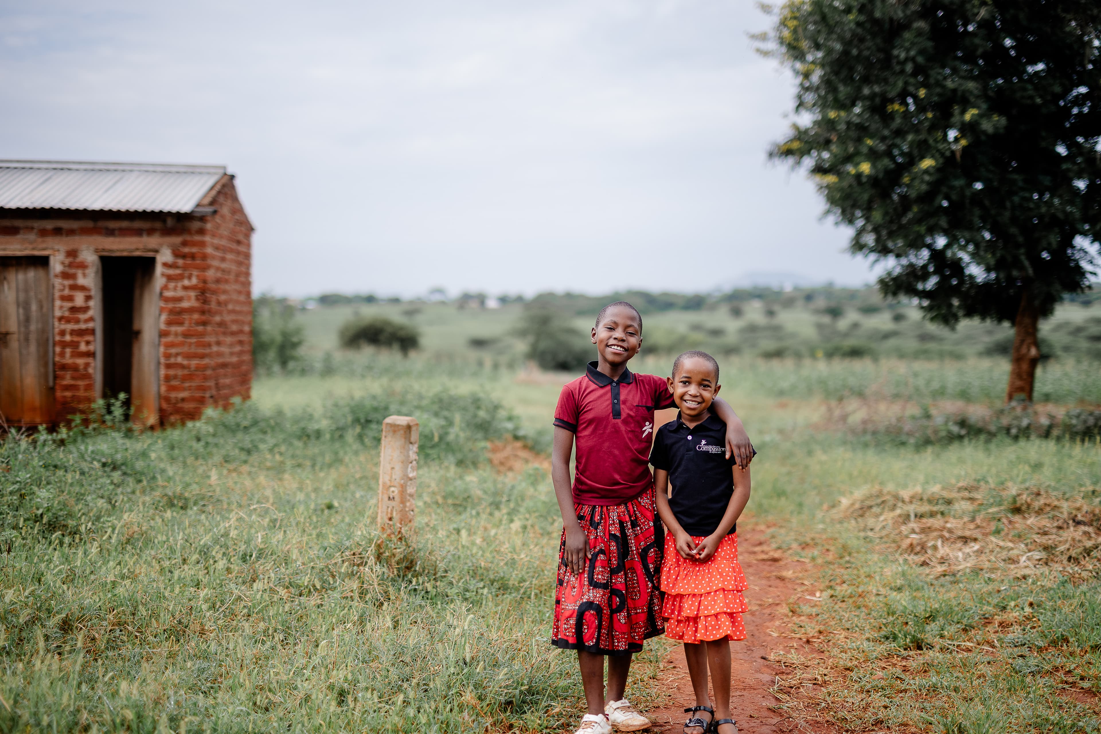 Two young girls stand outside on a red dirt path, smiling with their arms around each other, with grass and a brick building in the background.