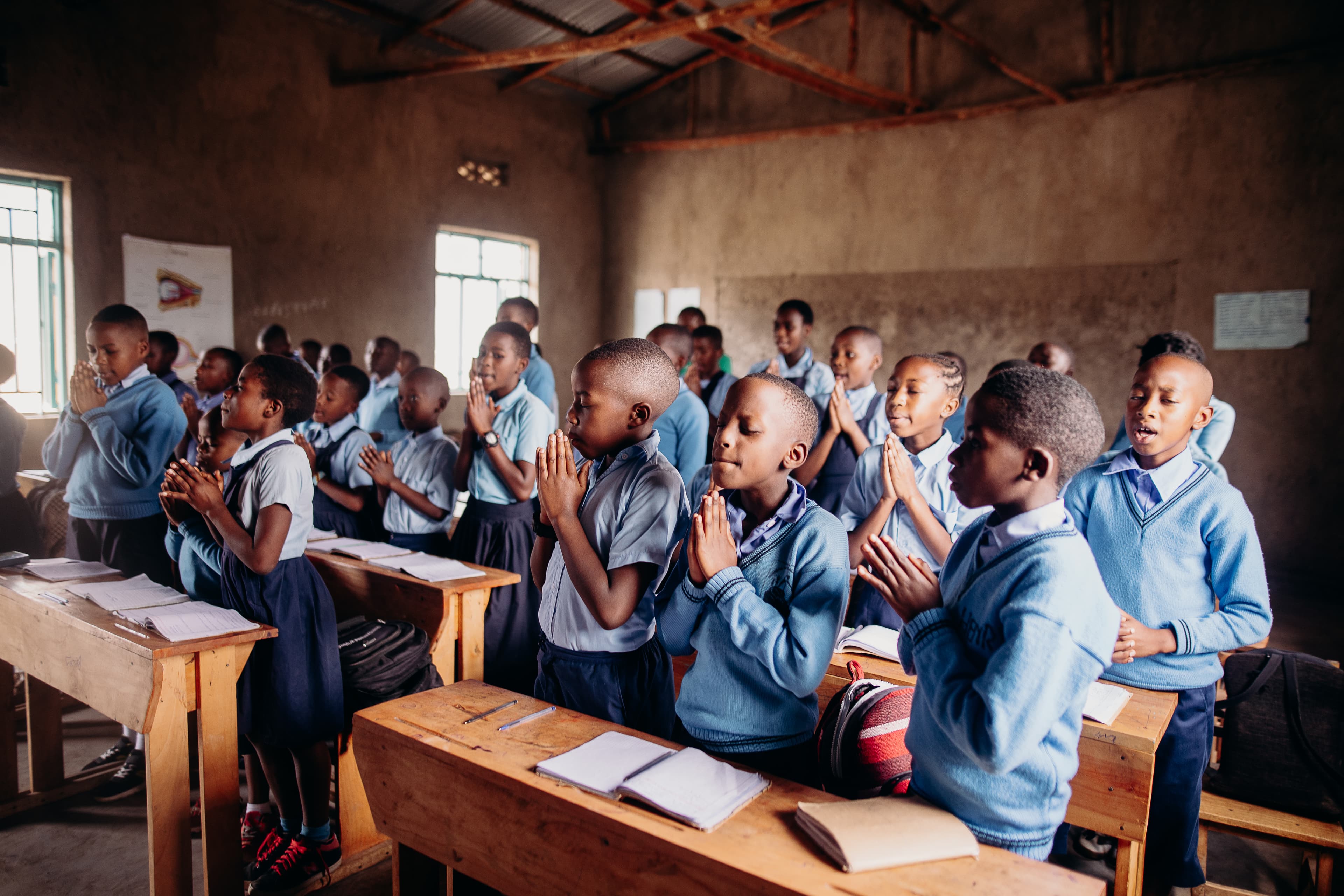 A classroom of children clasp their hands together while praying.
