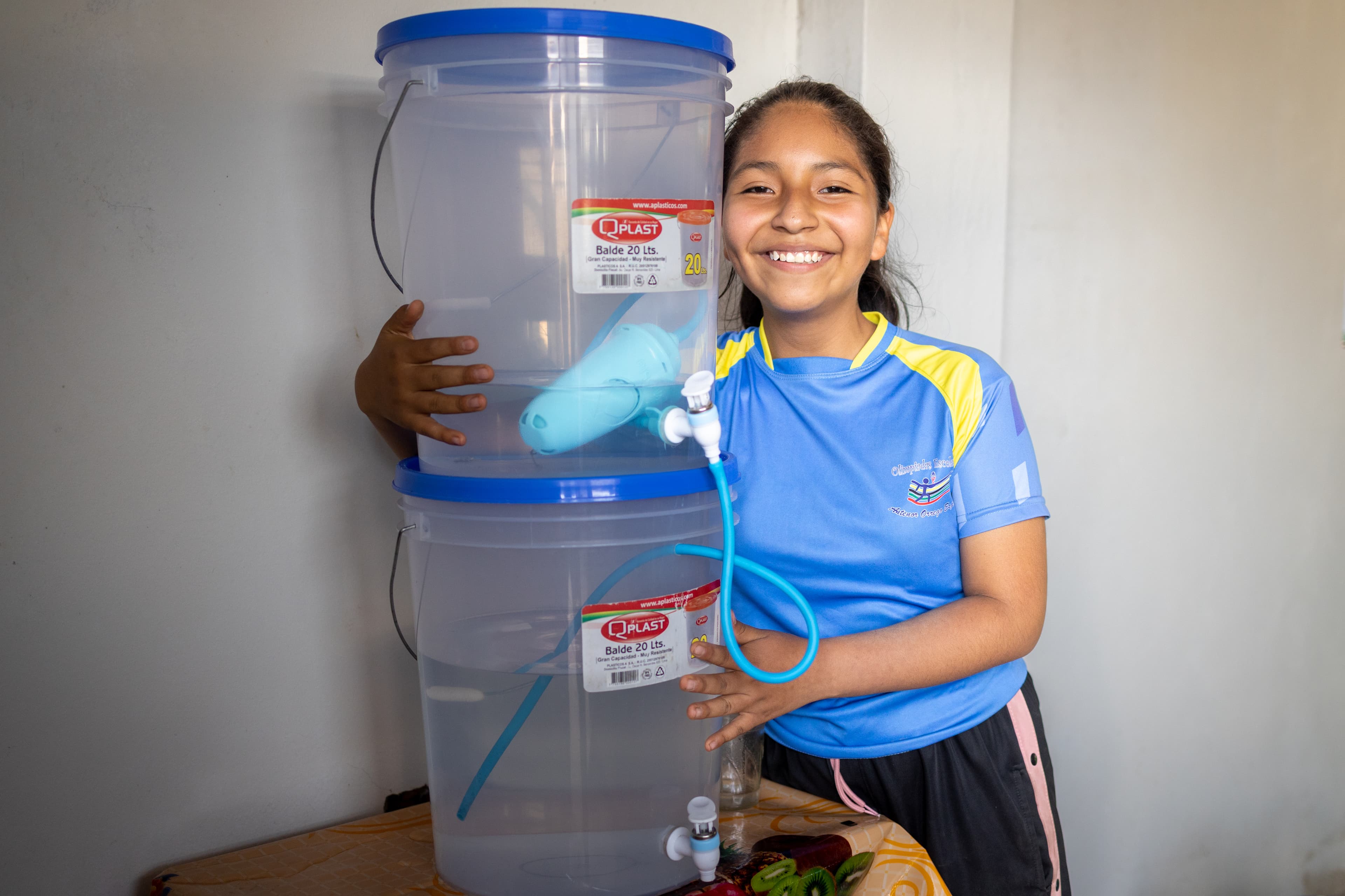Pre-teen girl wearing a blue and yellow shirt with black and pink shorts smiles while holding a large water filter.