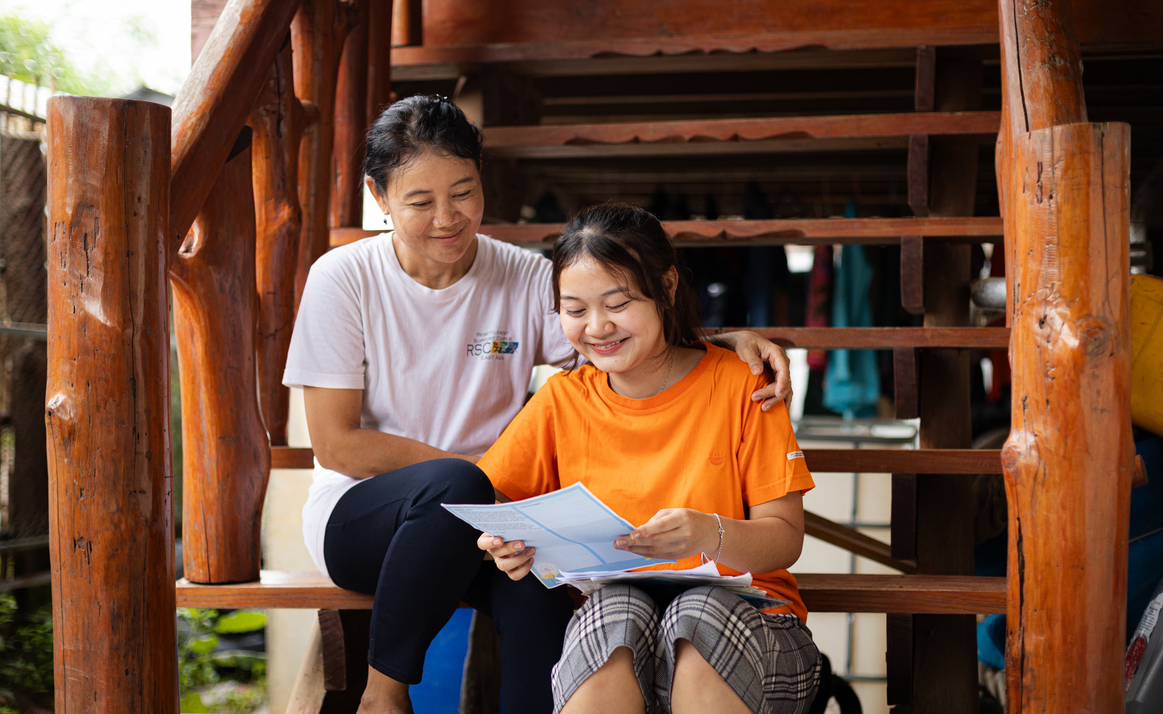 A teenage girl wearing an orange shirt and holding a letter sits on outdoor stairs with an older woman wearing a white shirt.
