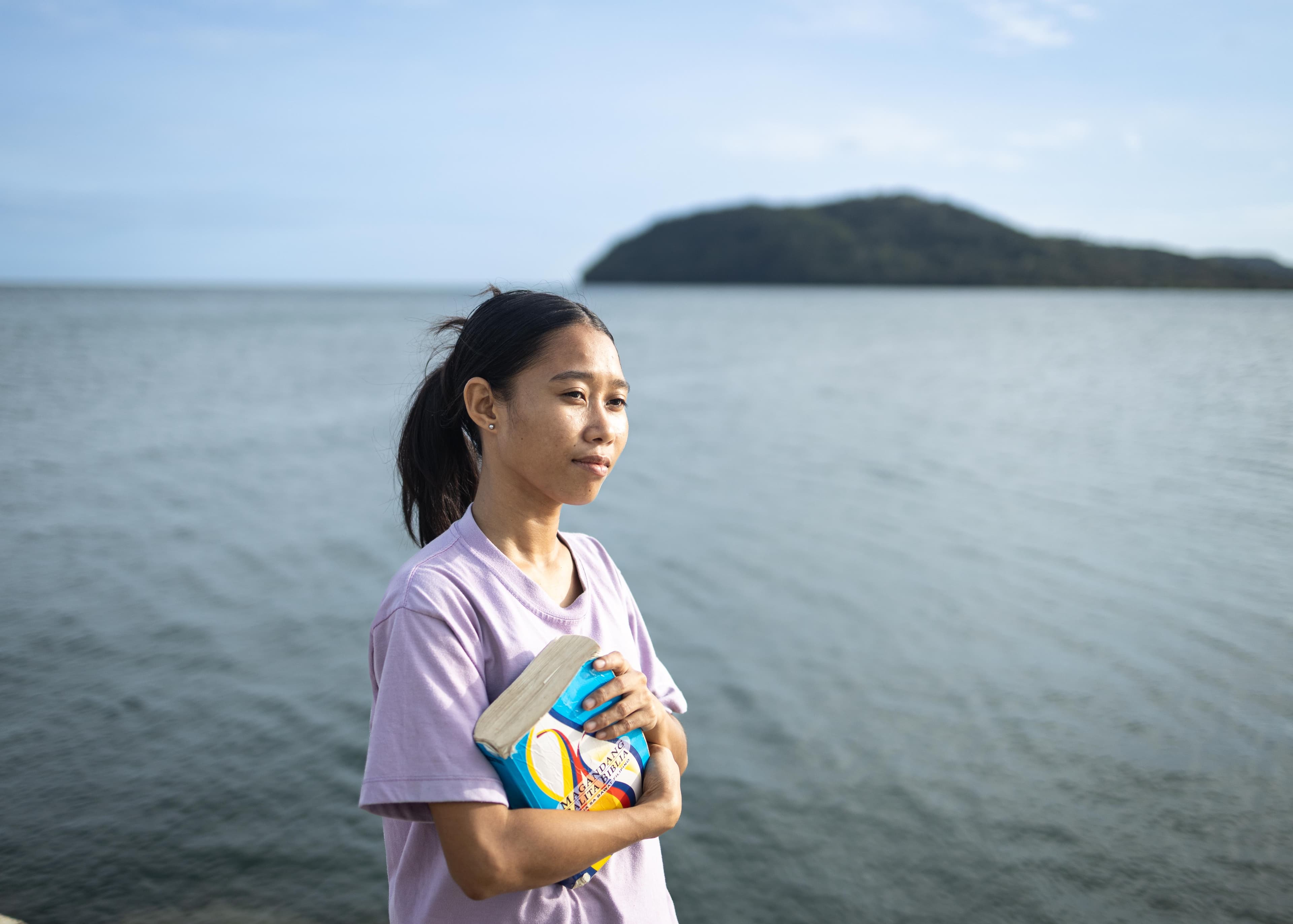 A young woman in a purple shirt stands holding her Bible in her arms as the sea flows behind her.