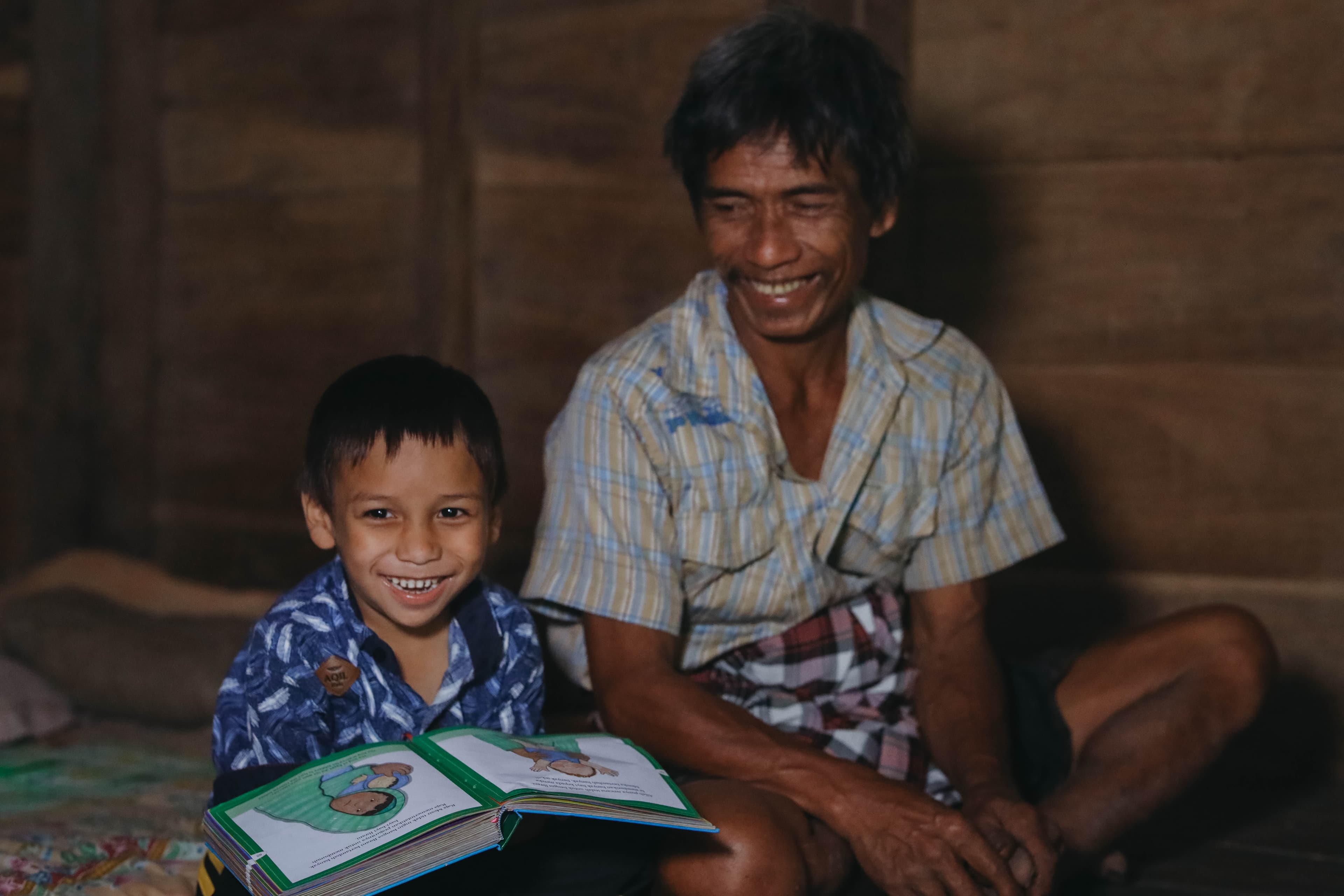A young boy is holding an open book and smiling as his father, wearing a yellow and blue shirt, smiles at him.