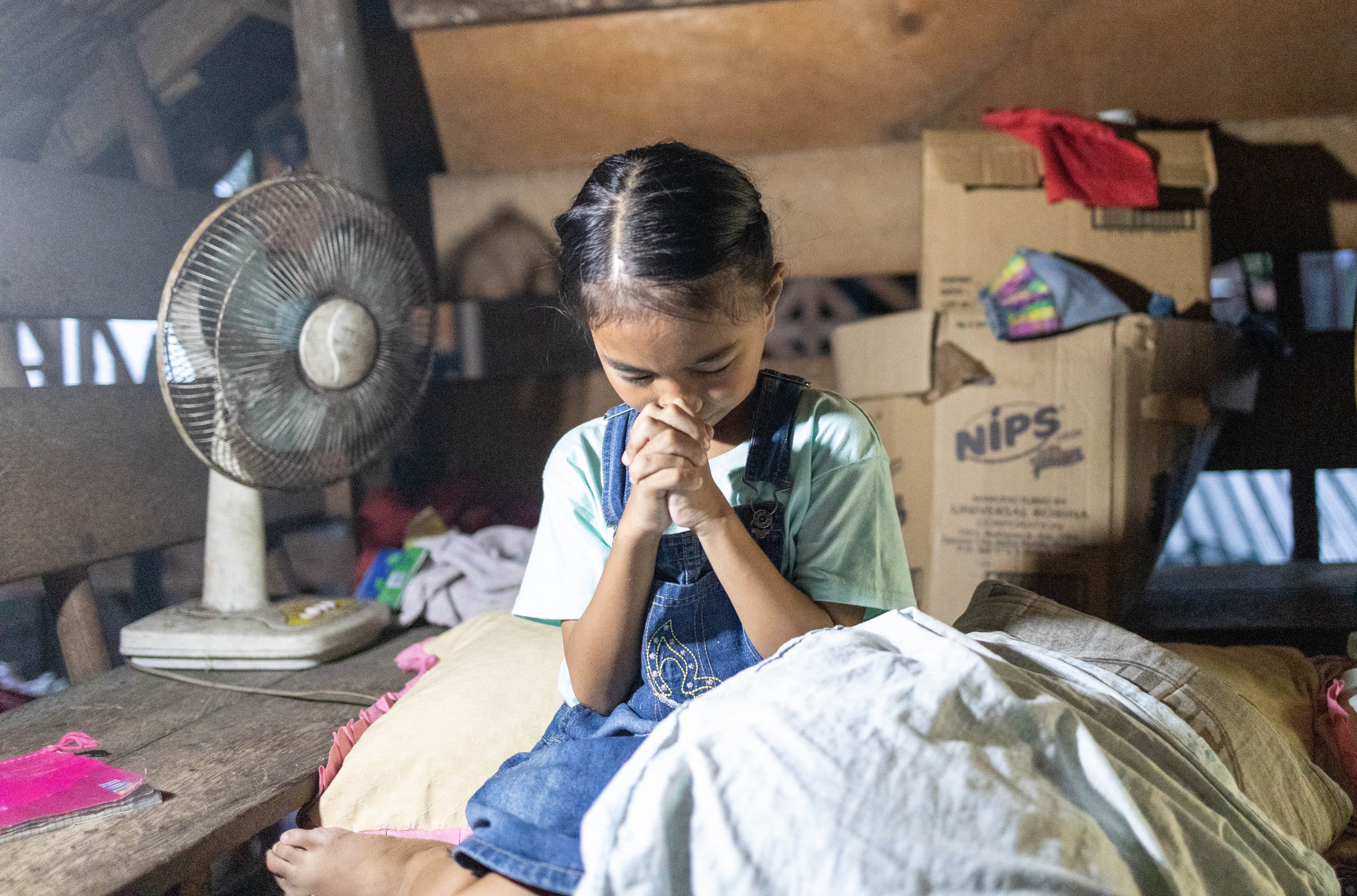 A young girl is sitting in her home with her head bowed praying as she rests her face on her hands.