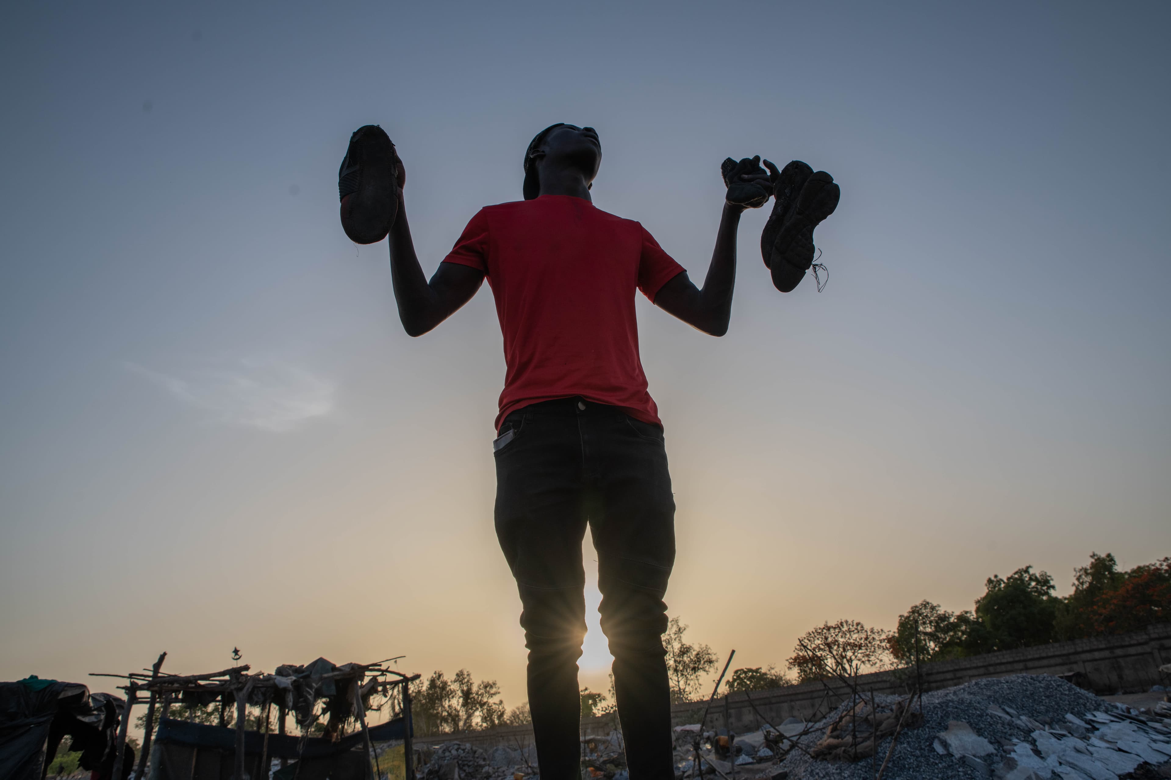 An African boy holds shoes in the air while the sun shines behind him.