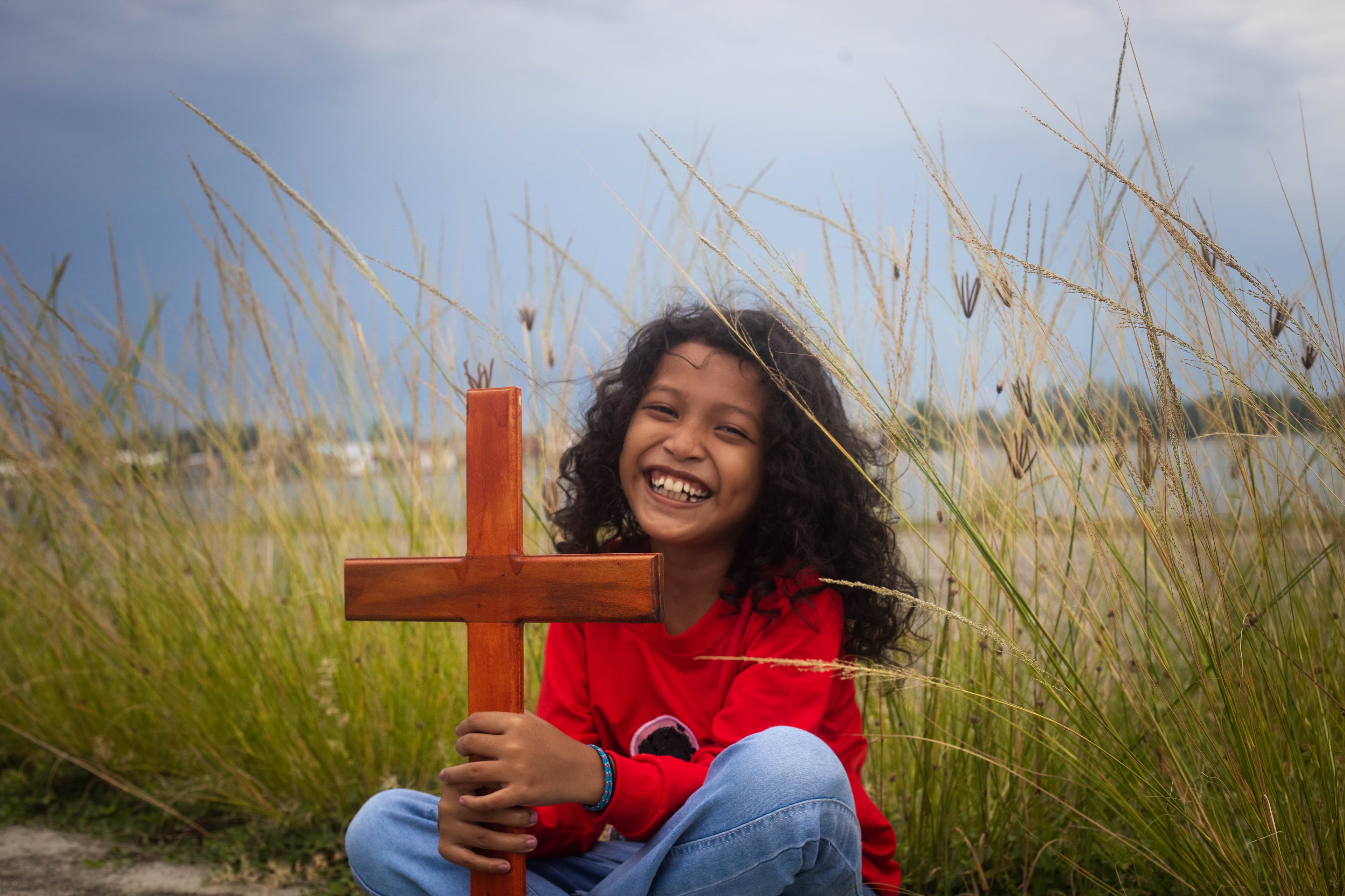 A young girl wearing a red shirt sits in a field while holding a wooden cross and smiling.