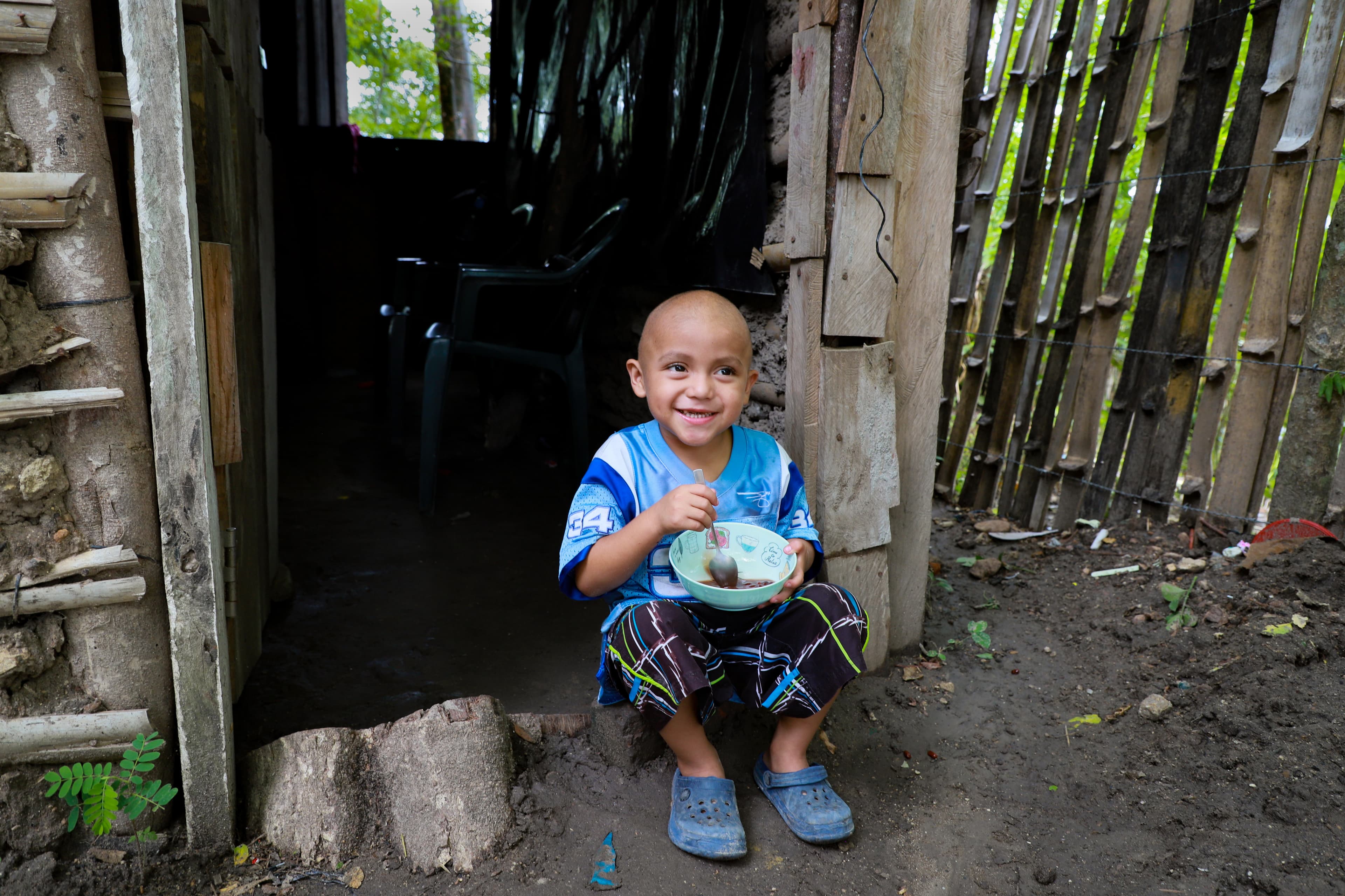 A young boy is sitting outside his home holding a spoon and a bowl of food smiling.