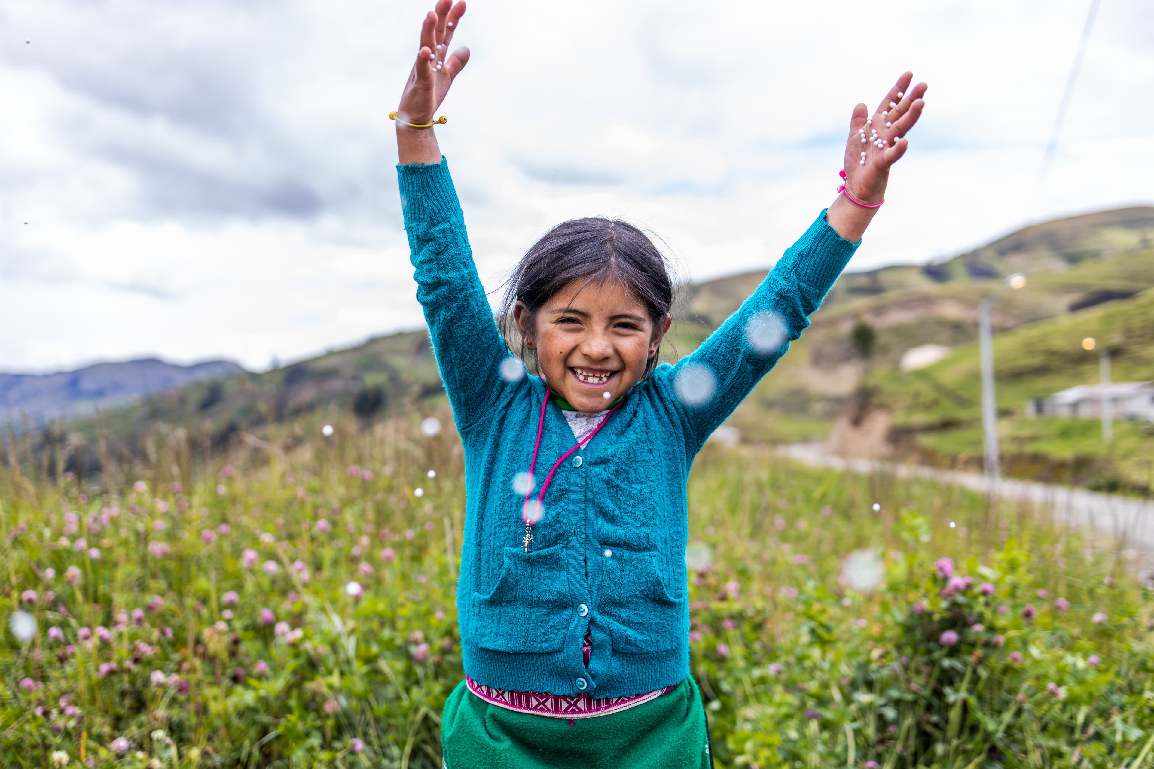 A young girl is standing in a field of wild flowers and jumping with her arms in the air.