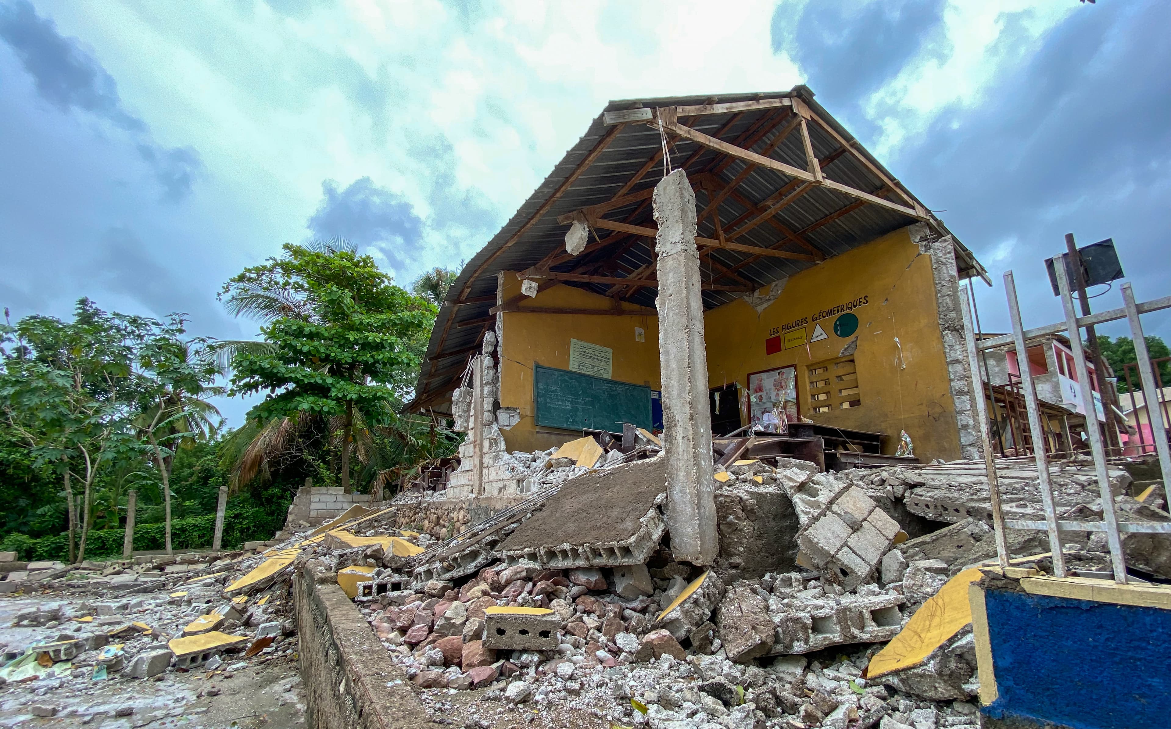 A roof and two walls surrounded by rumble due to an earthquake in front of green trees and a cloudy sky.