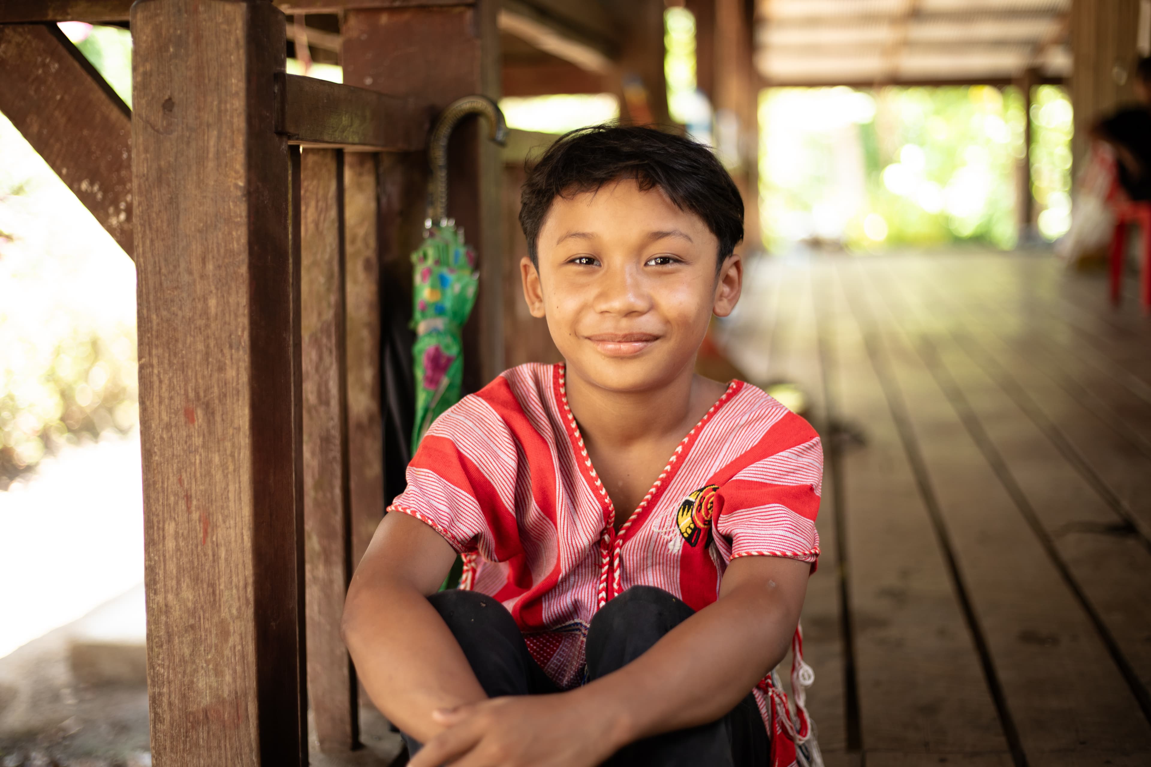 A boy dressed in a pink and traditional shirt sits on a wooden porch with his knees to his chest, smiling.