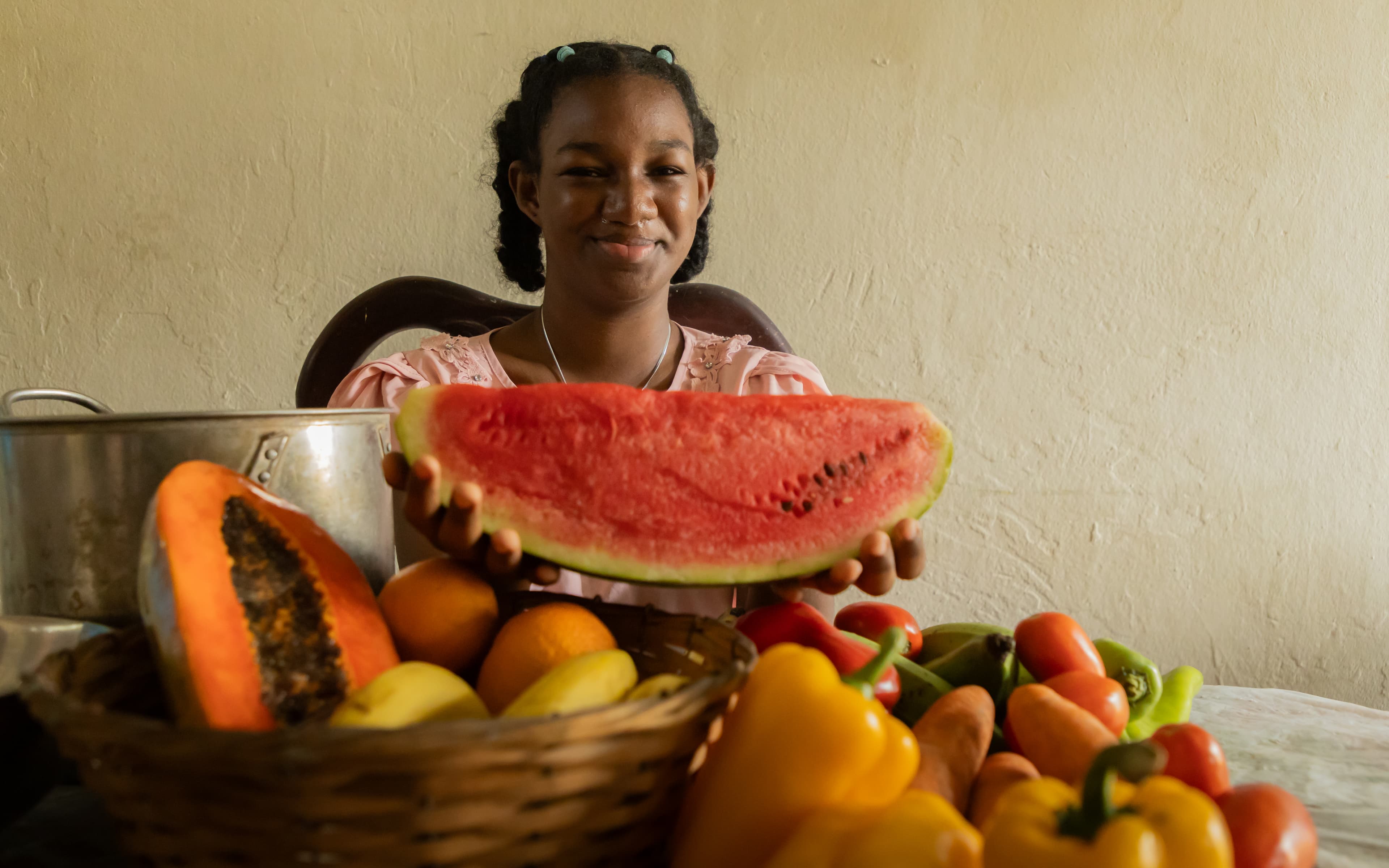 A girl sits at her kitchen table holding a watermelon in her hands with fruits and vegetables laid out in front of her.