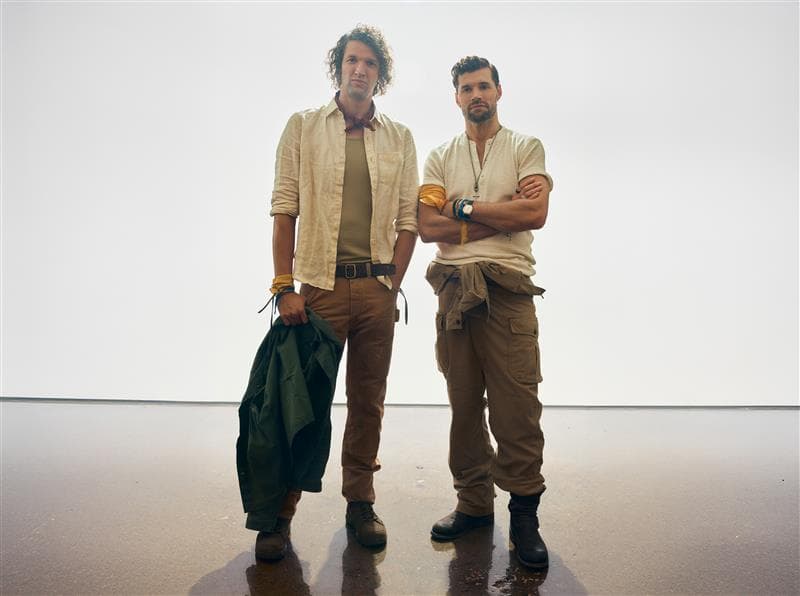 Two men stand next to each other, one with his arms crossed and one holding a green jacket, in front of a white wall.