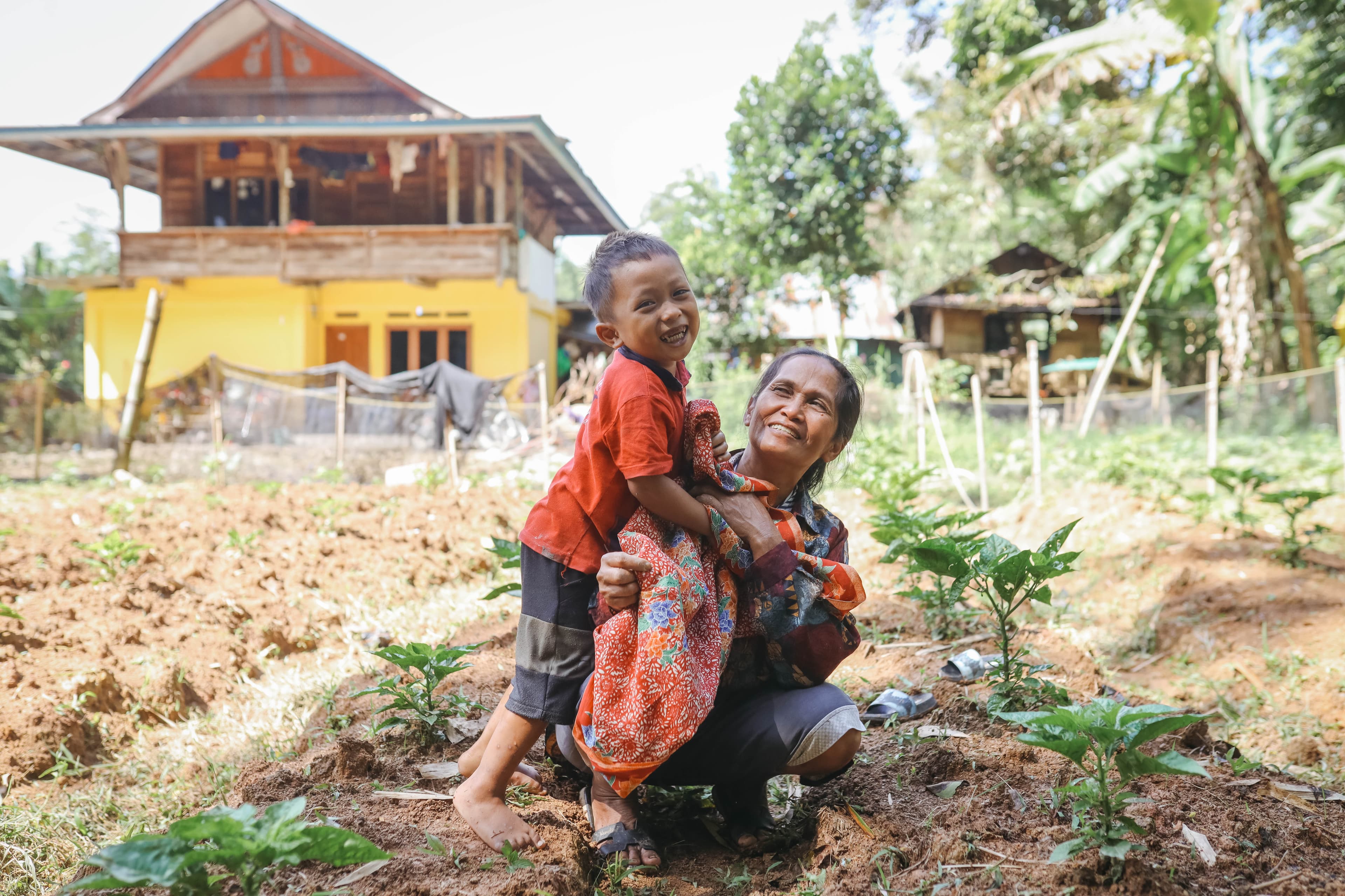 Young child and mother smile and kneel in a field with seedlings around them.