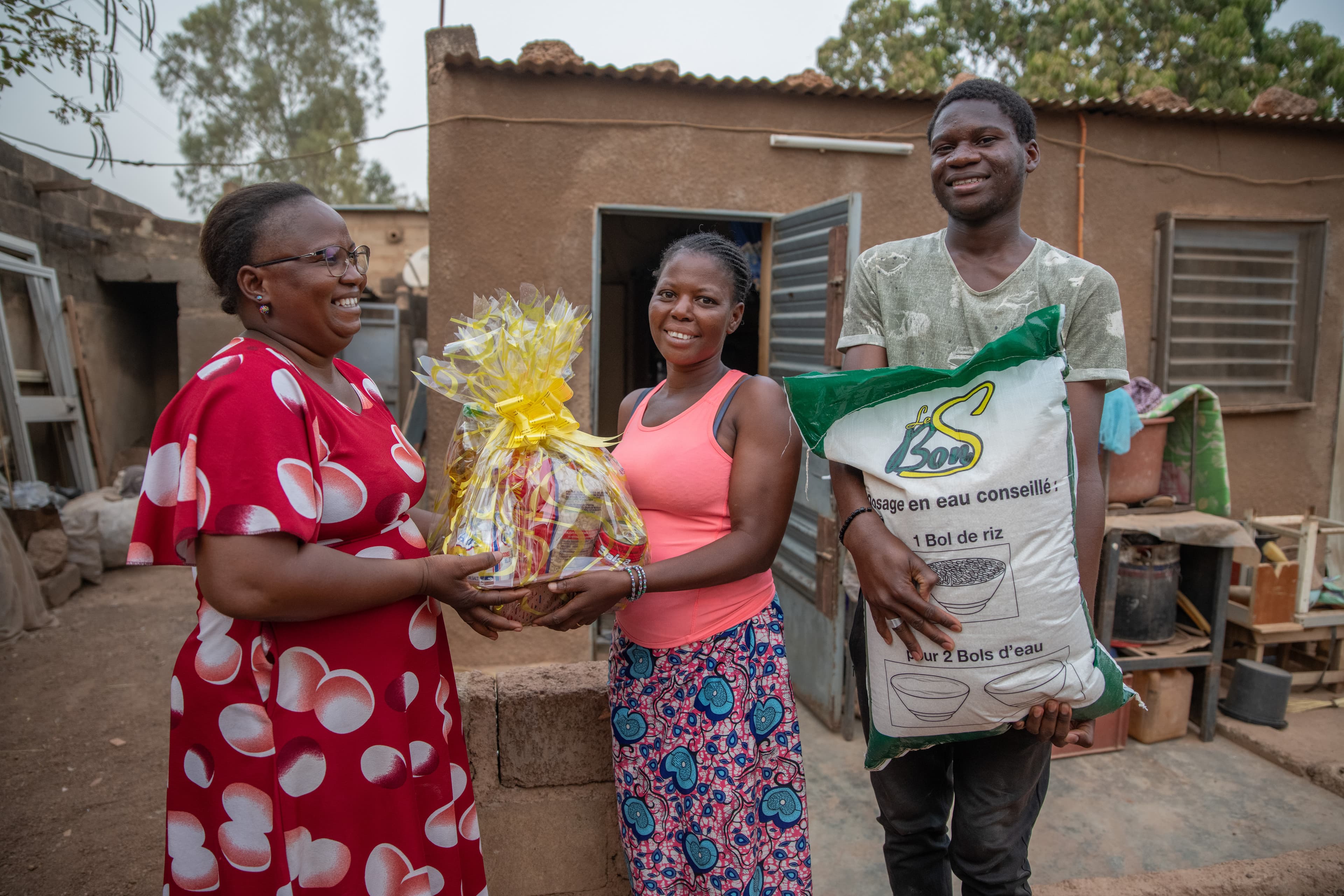An older African woman gives a basket of food to an African couple as they all smile for the camera.