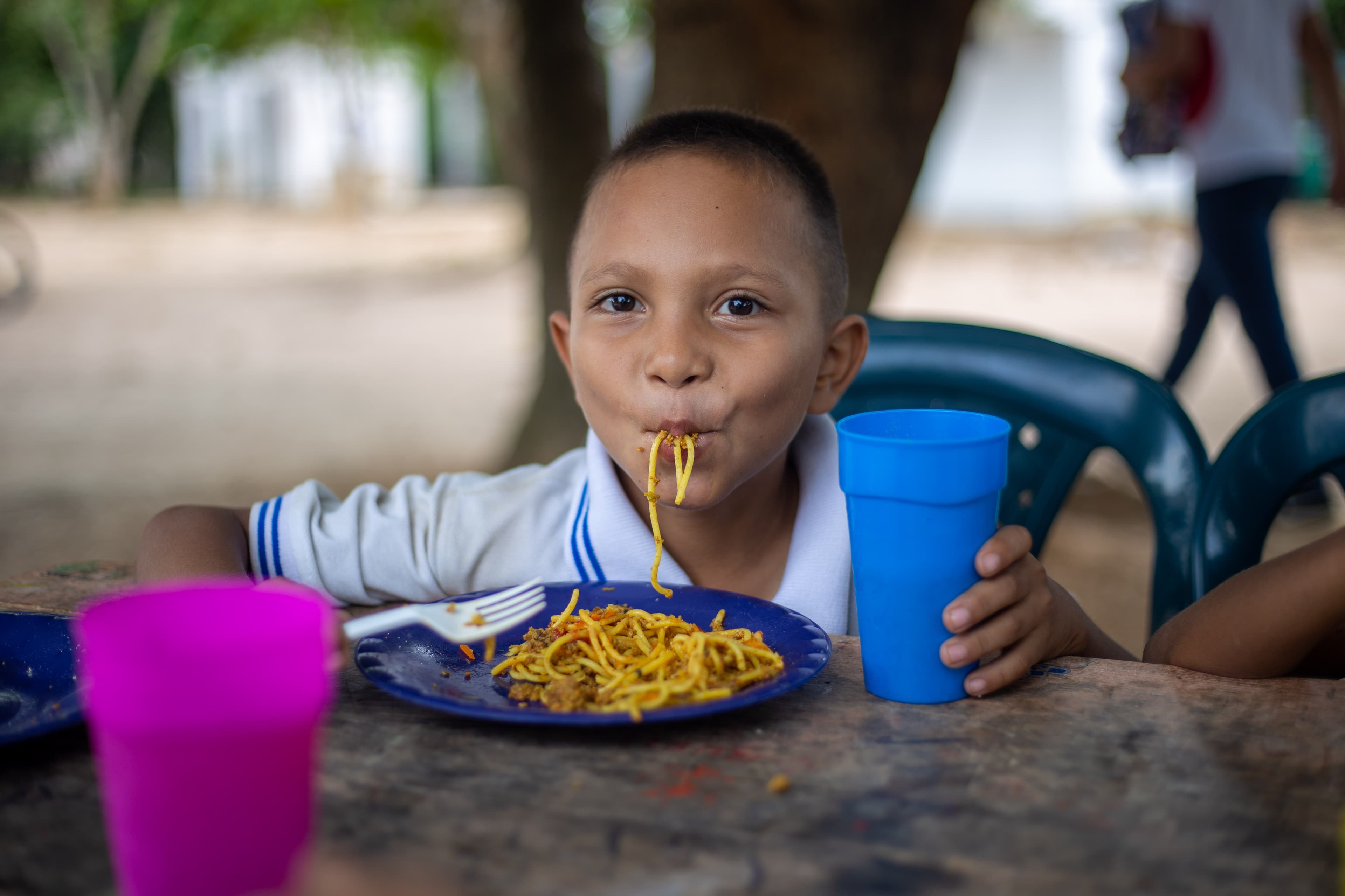 A young boy is sitting at a table eating a plate of spaghetti and holding a cup.
