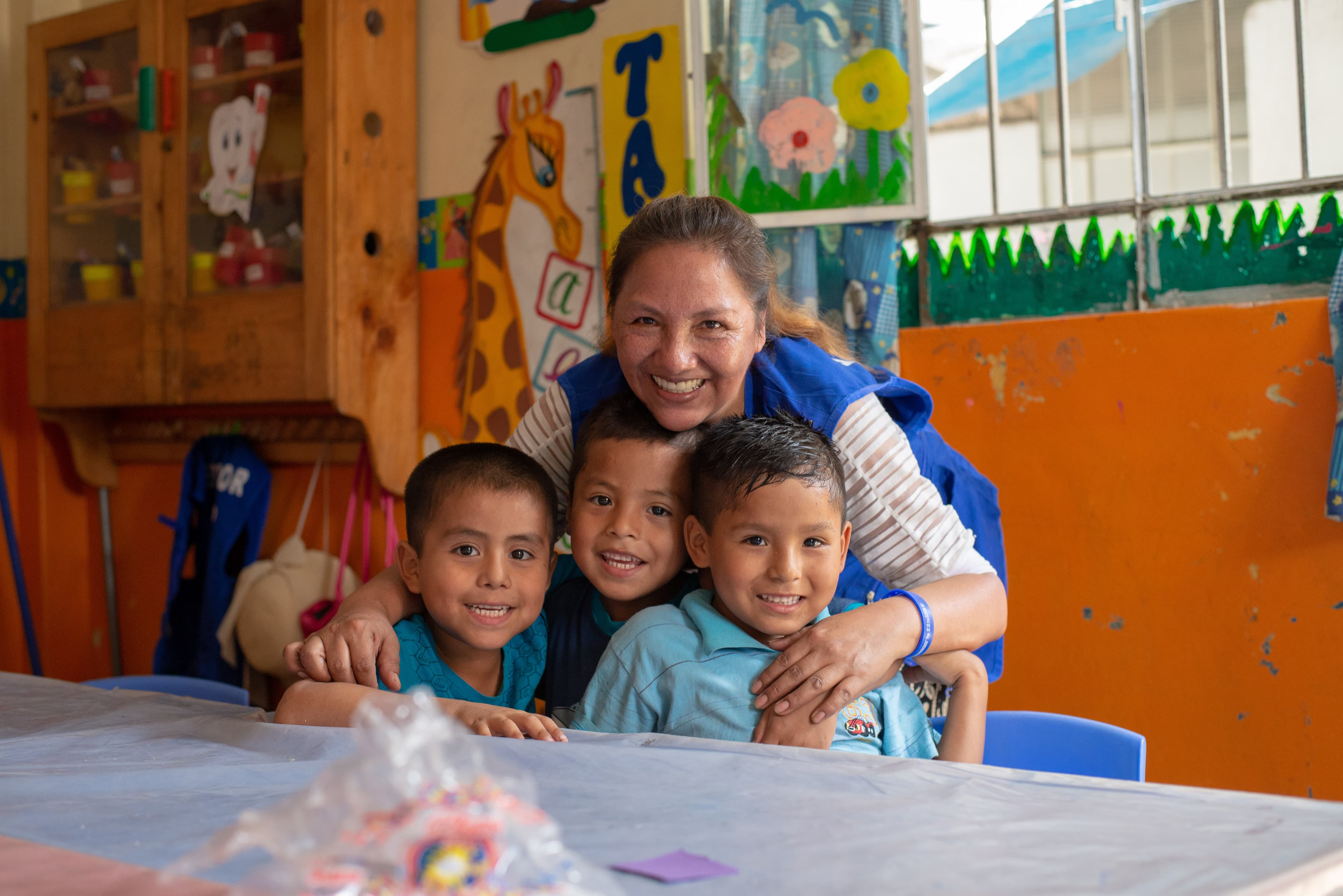 Woman hugs three smiling boys sitting at a classroom table
