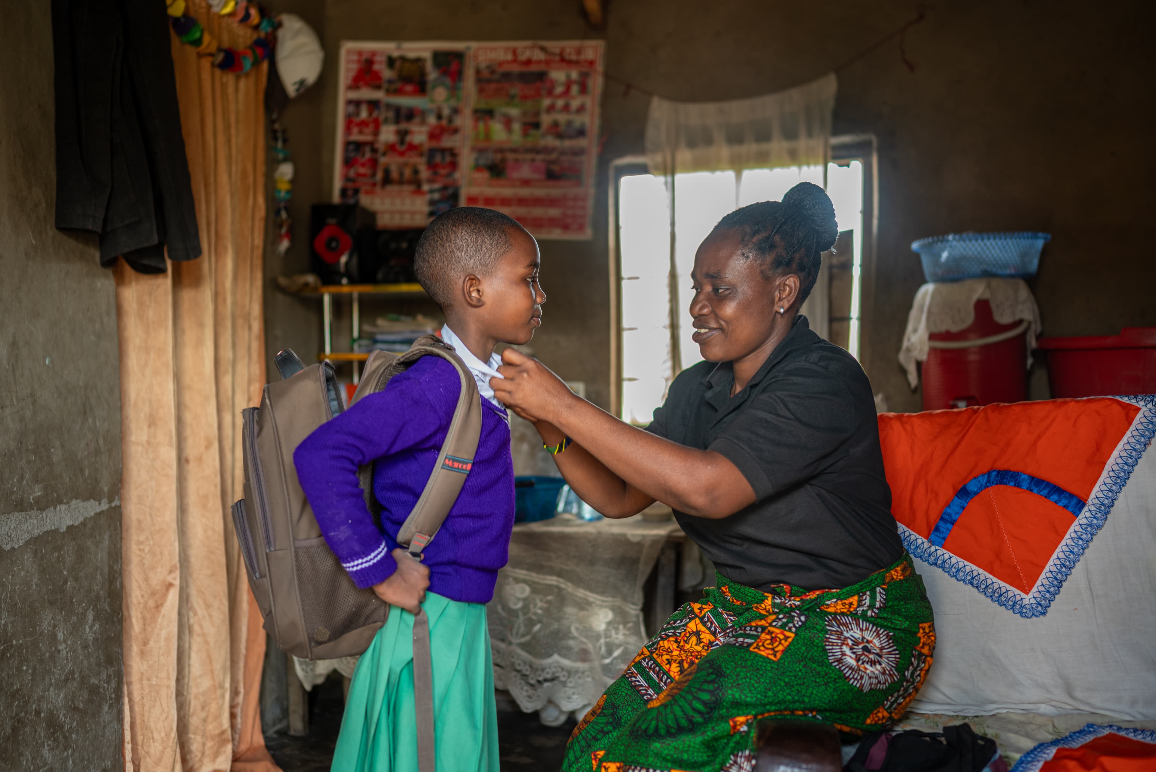 An African mom helps her daughter put on her school uniform while smiling.