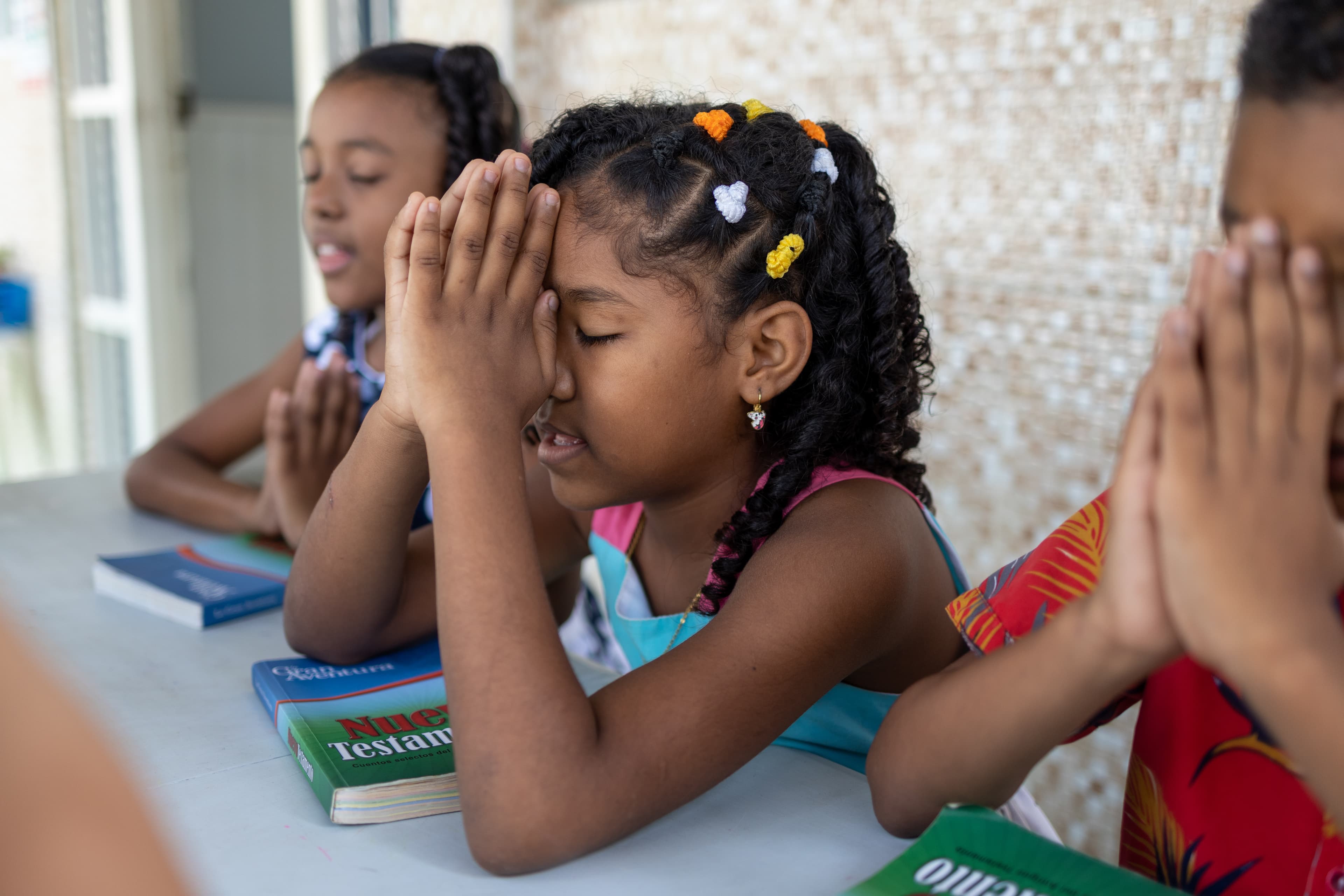 Young Colombian girl in a pink and blue dress sits at a table, praying with other children.