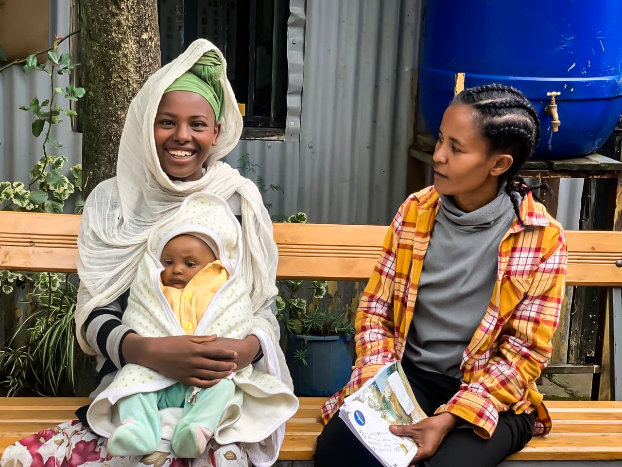 A Compassion Survival staff member is wearing a plaid shirt, a gray turtleneck, and black pants. A mother is wearing a white shawl and has a green head-covering and her baby, Edlawit, is wearing yellow and green and is wrapped in a blanket.