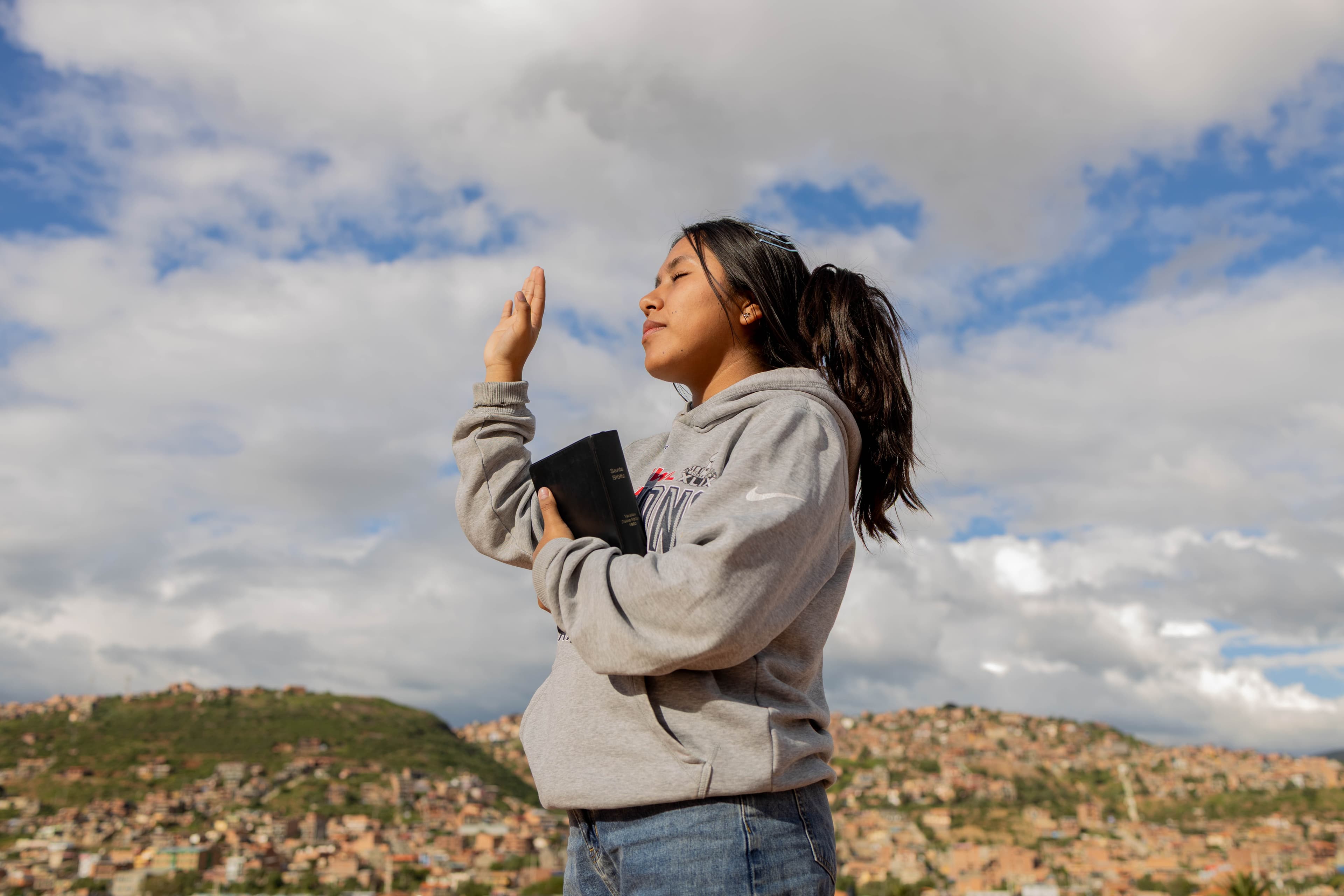 A young girl holds her hand up in prayer with her eyes closed and her bible against her chest.