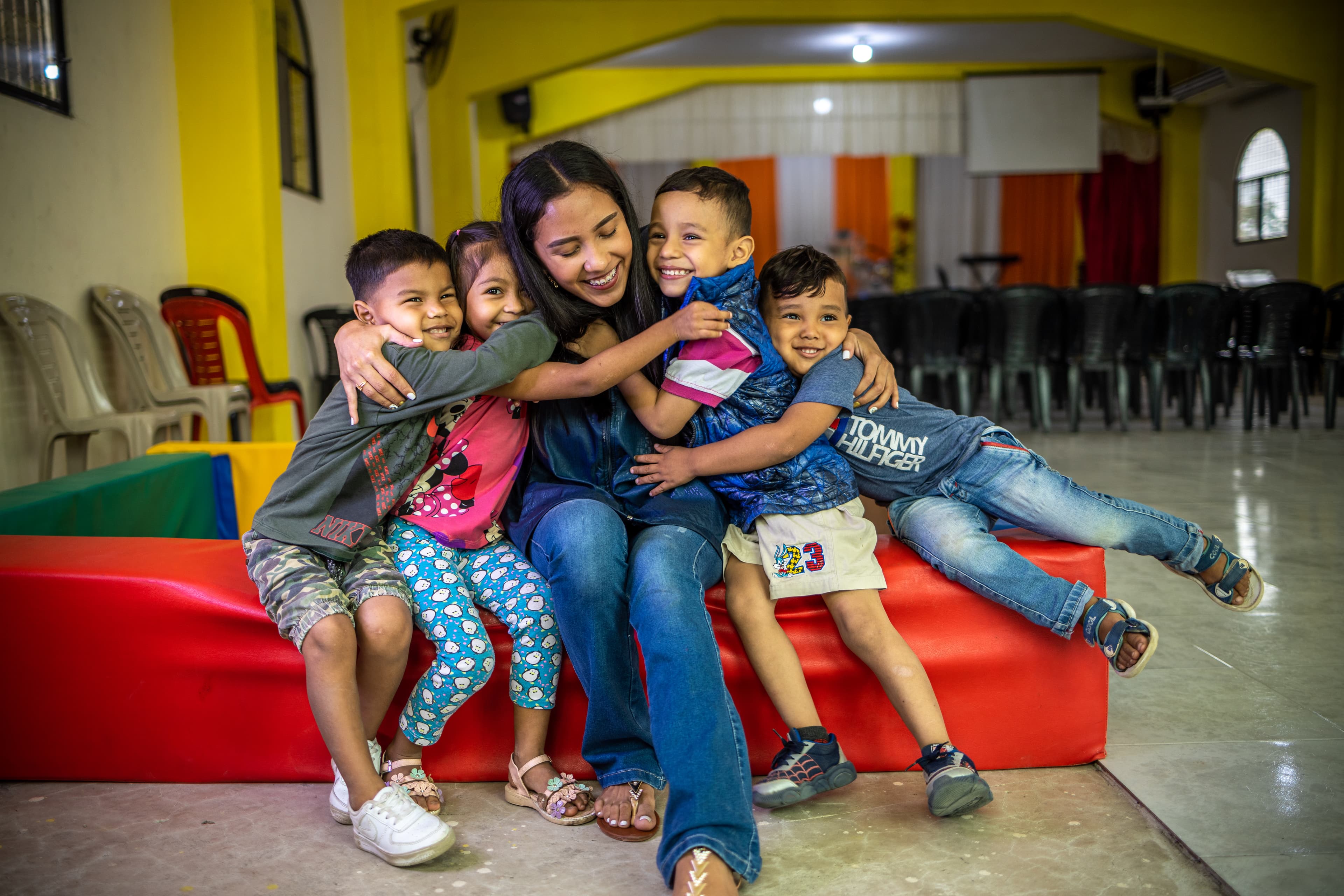 A woman wearing a blue shirt and jeans is sitting inside of a church. There are four children smiling and hugging her.