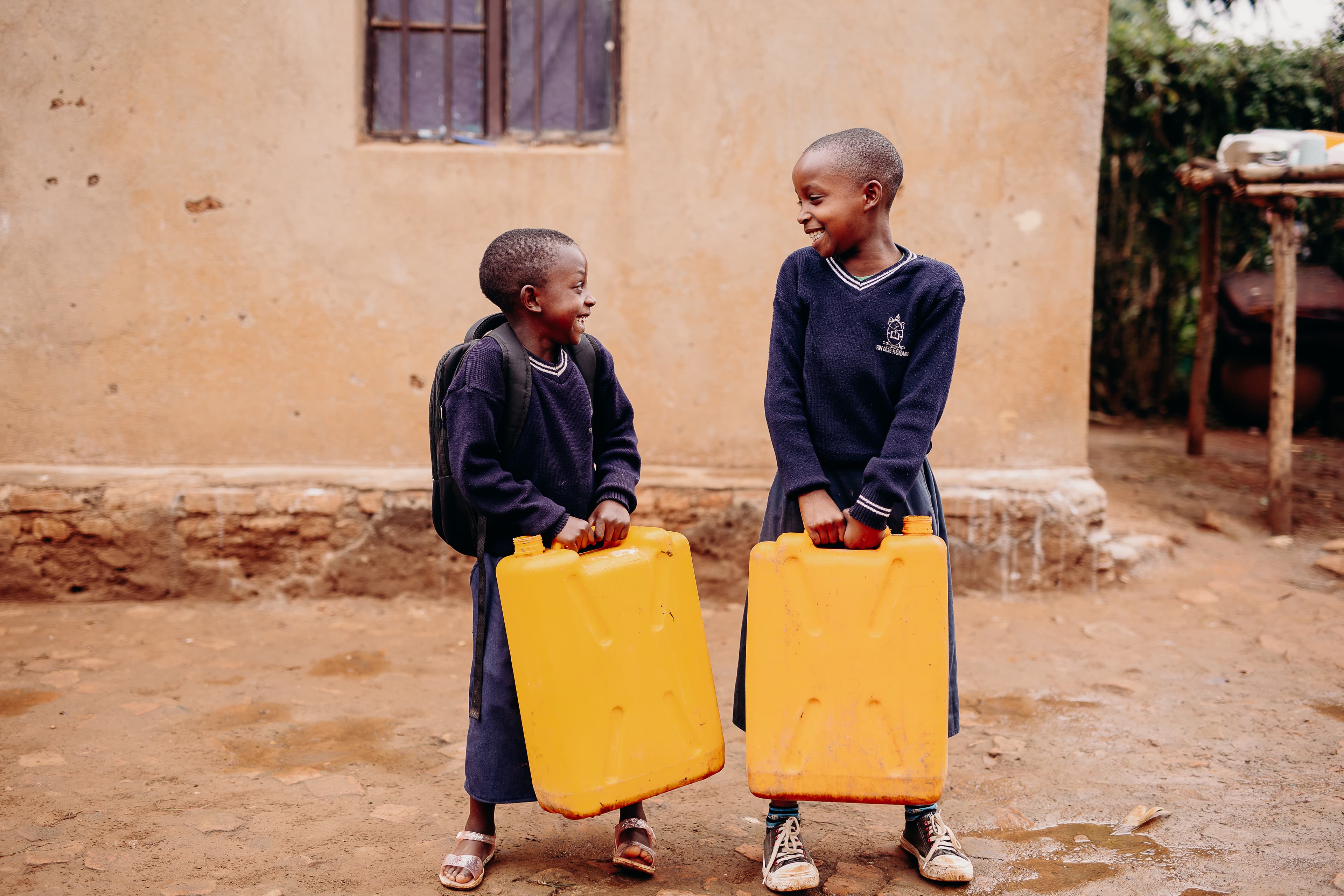 Two Rwandan boys in school uniforms look at each other while holding large, yellow water jugs.