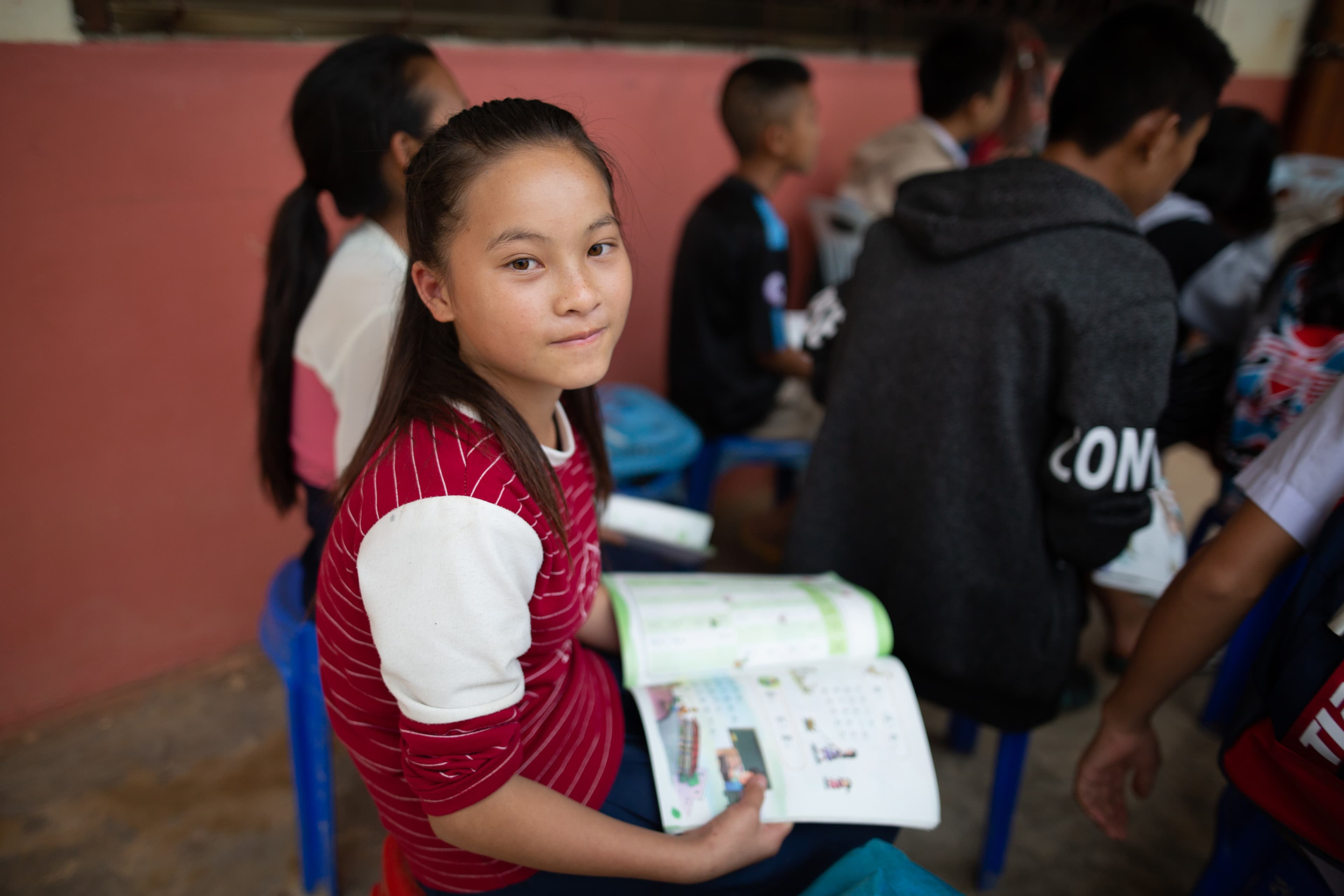 Young girl sits in a classroom chair surrounded by students with a book in her lap.