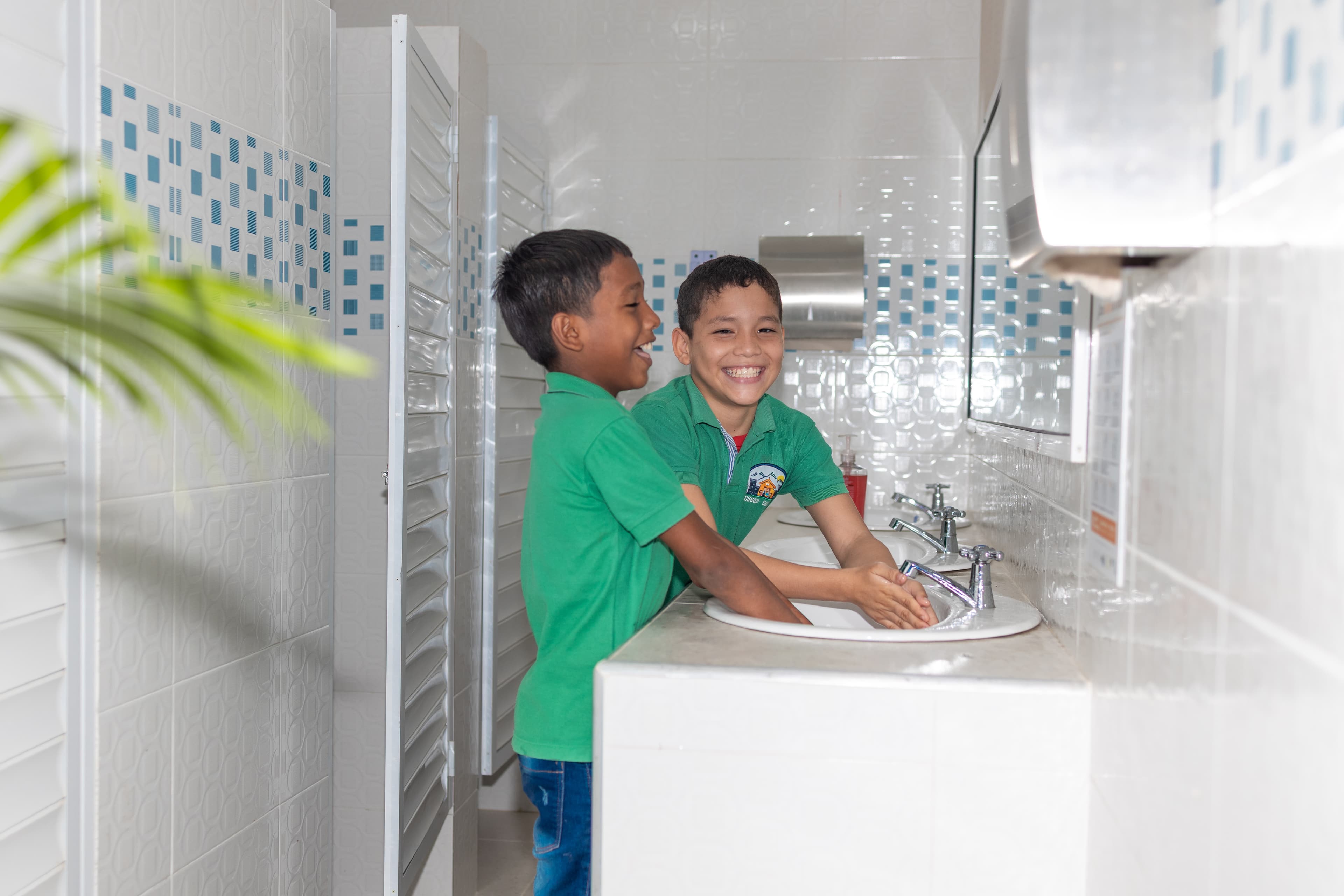 Two Colombian boys wearing green shirts wash their hands in a sink while laughing and smiling.