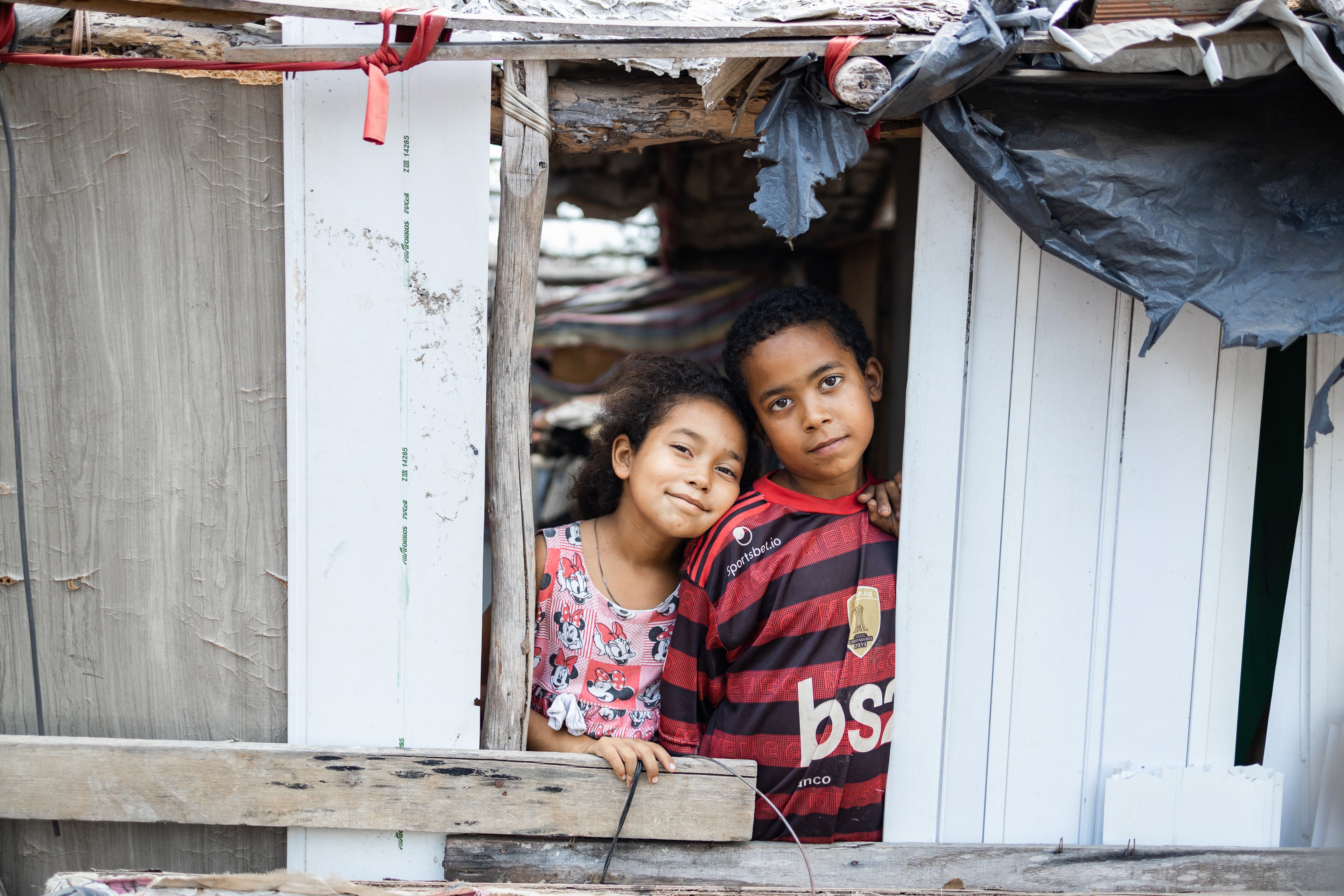 Two young children stand inside of a window and smile.